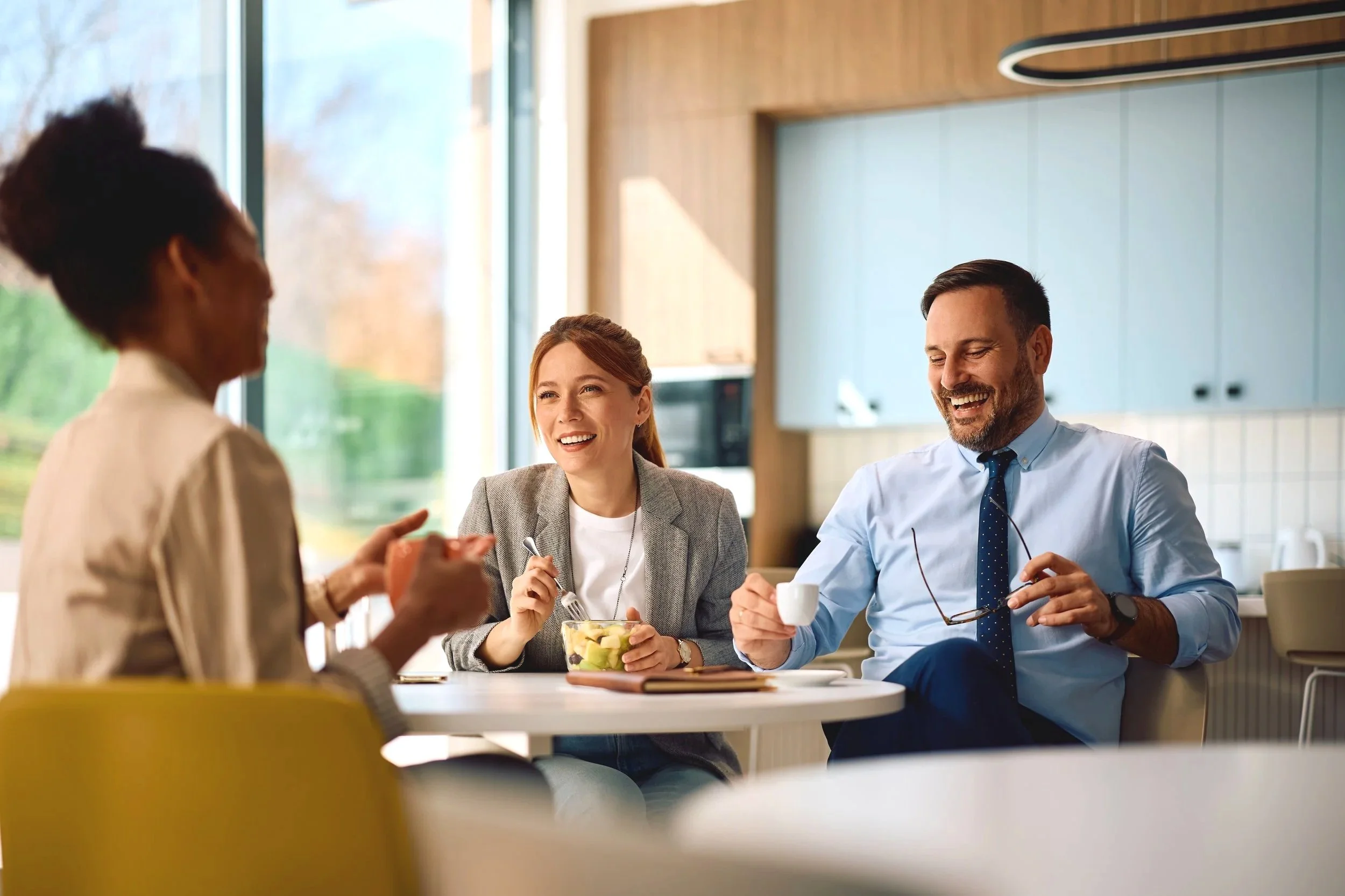 Business professionals having a meeting and enjoying coffee in a modern office break room with large windows and natural light.