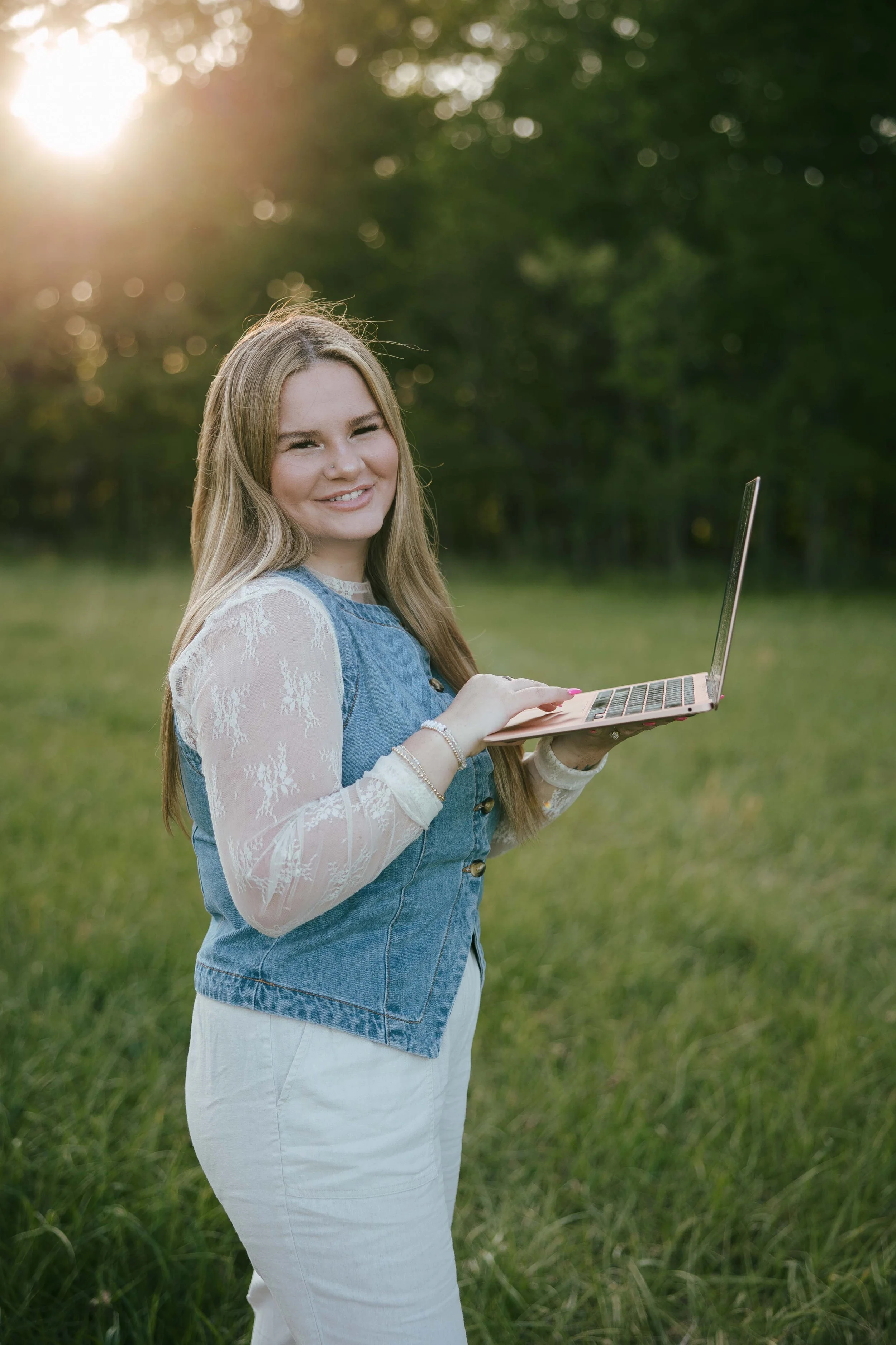 A young woman with long blonde hair, wearing a white lace long sleeve shirt under a blue denim vest and white pants, is smiling and holding an open laptop outdoors in a grassy field during sunset with green trees in the background.
