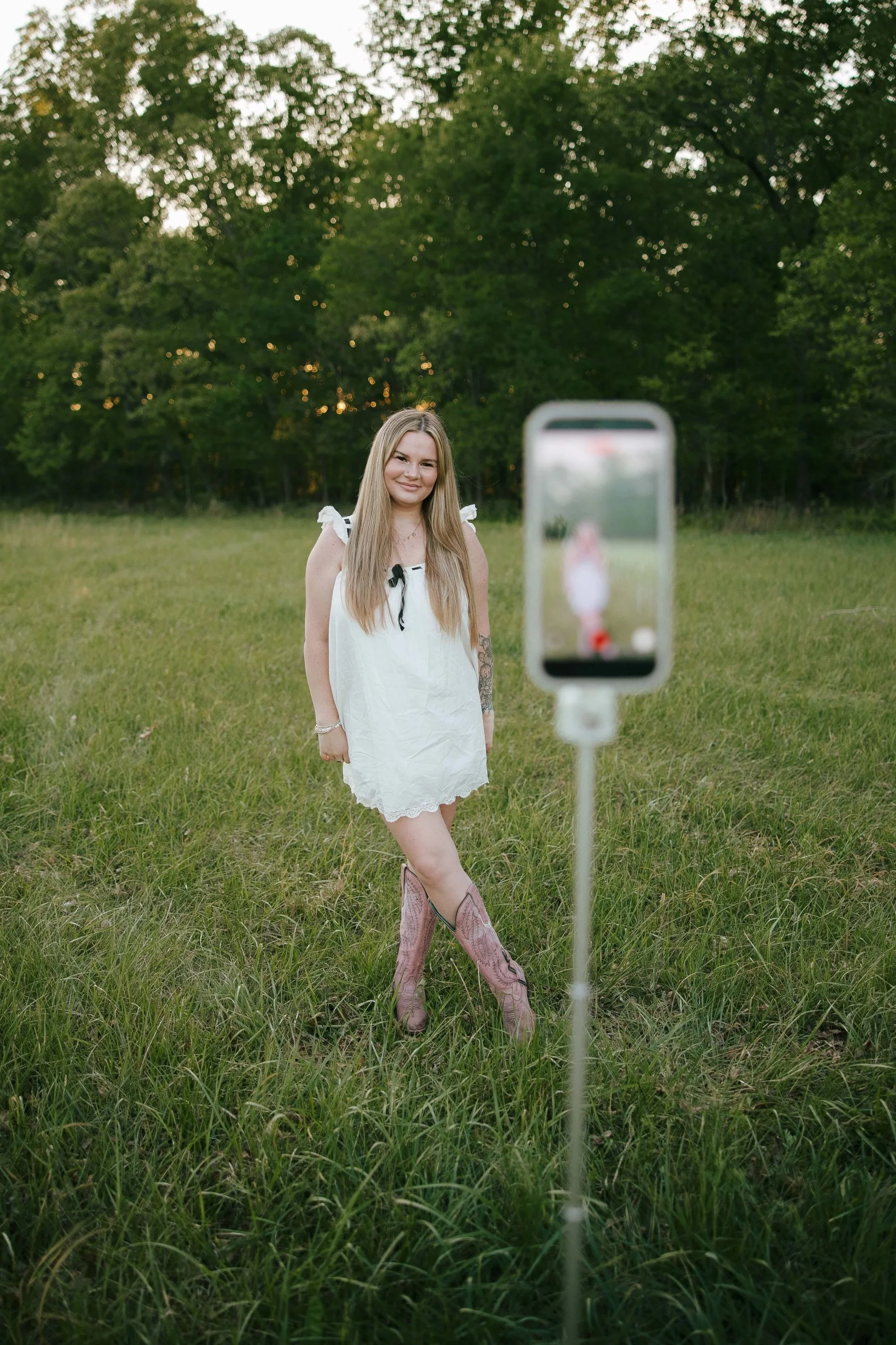 Young woman in a white dress taking a selfie in a grassy outdoor field with trees in the background creating Instagram reels for local businesses