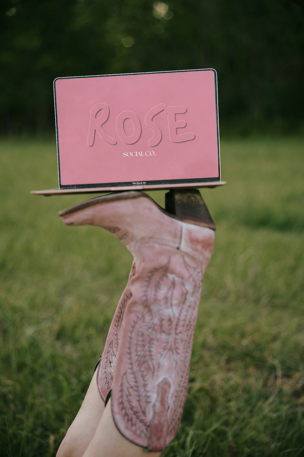 A person lays on their back with legs raised and intertwined, wearing cowboy-style boots with intricate stitching, holding a pink laptop displaying a pink background and the words 'ROSE SOCIAL CO.' in large letters.