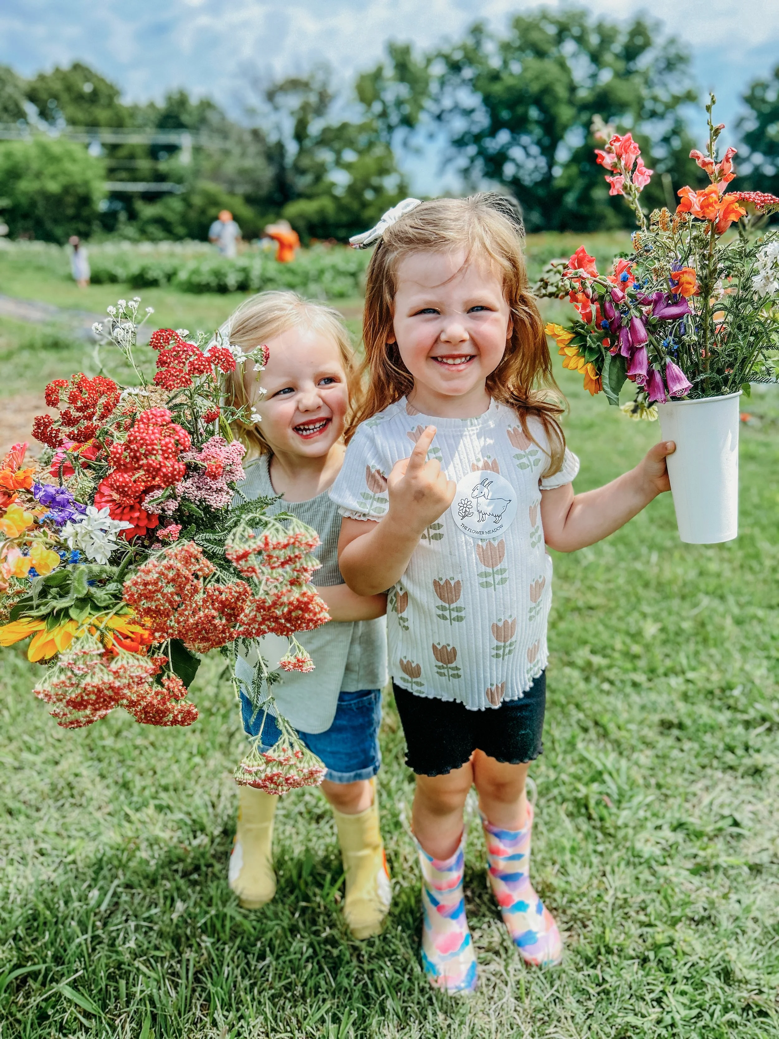 Two young girls standing outdoors on a grassy field amid colorful flowers, smiling at the camera. One girl is holding a white bucket with pink, purple, and orange flowers; the other girl stands behind her at The Flower Meadow You Pick Flower Farm