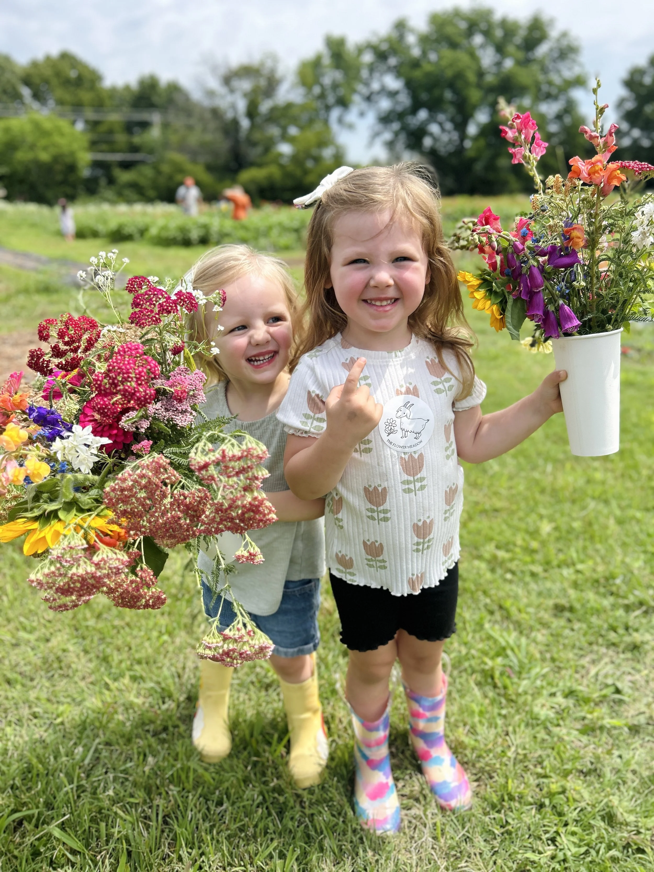 Two young girls with blonde hair in a field holding colorful flower arrangements,  at The Flower Meadow Flower Farm in Coweta, Ok and smiling.