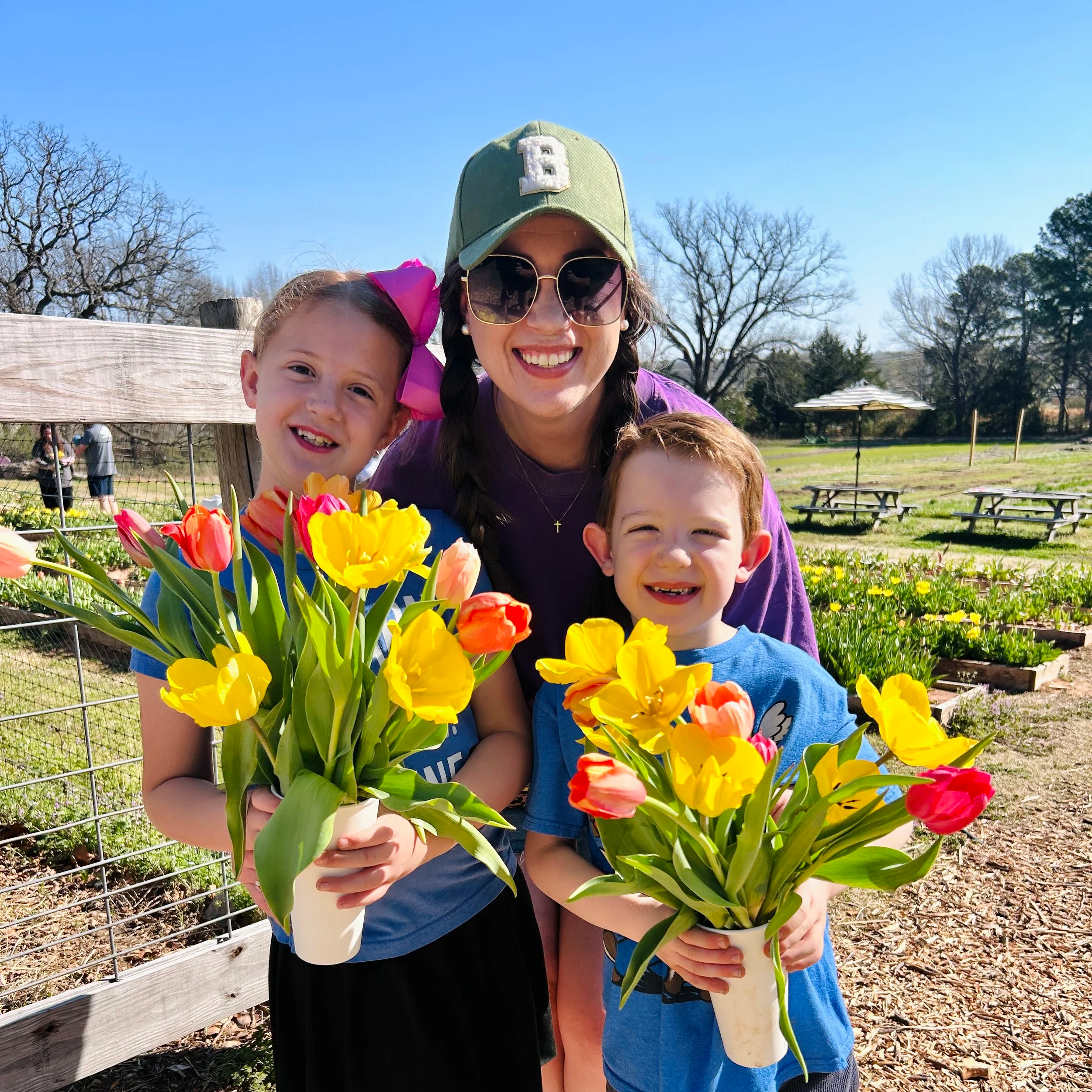 Smiling woman with sunglasses and a green cap holding two children at The Flower Meadow Flower Farm and Barnyard Petting in Coweta, Ok with tulips, trees, and a picnic table in the background.