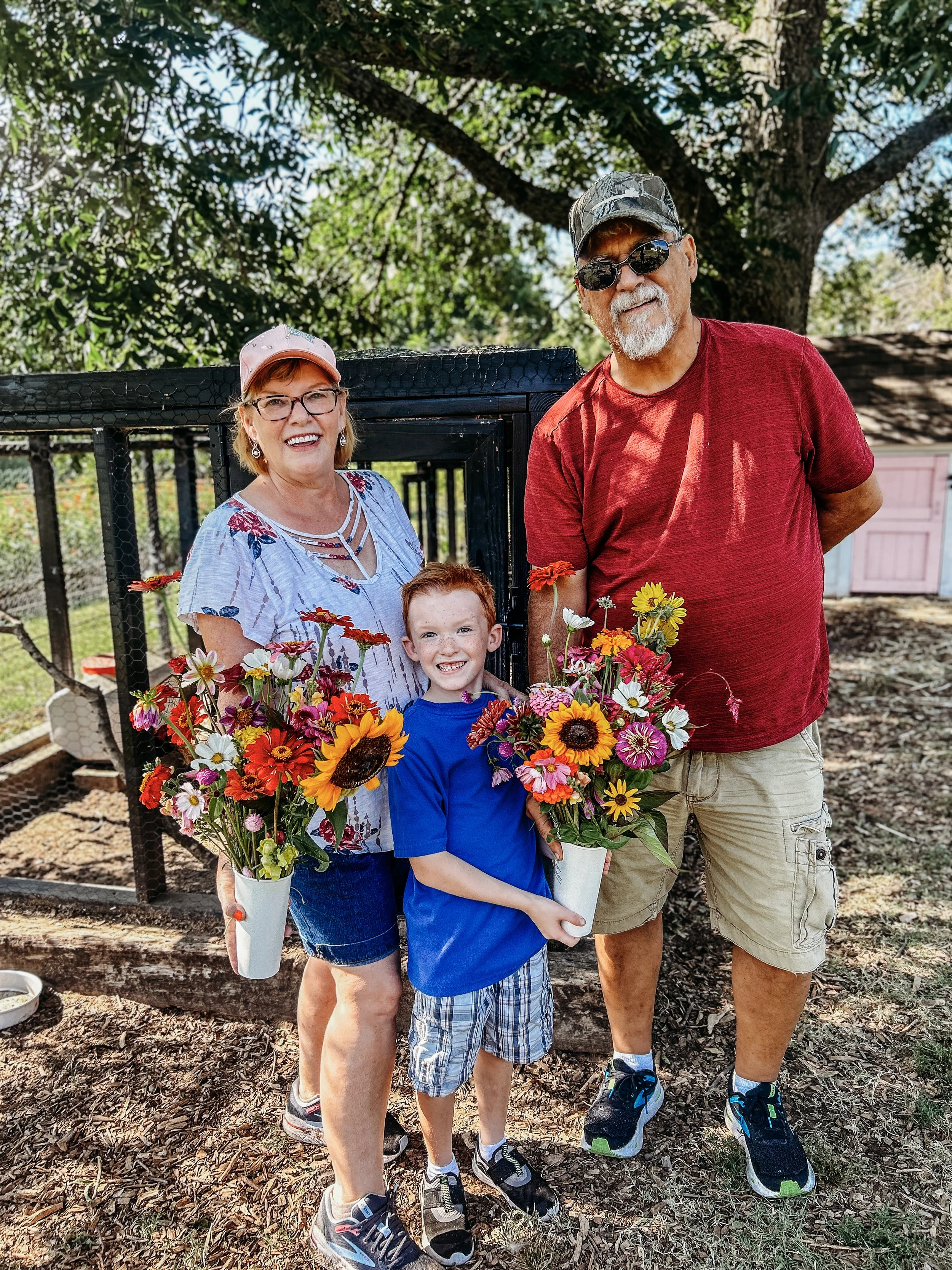 A family of three standing outdoors, holding colorful flower bouquets, smiling at the camera. The woman wears glasses, a floral shirt, and a pink cap. The young boy wears a blue shirt and plaid shorts. The man wears sunglasses, a red T-shirt, and khaki shorts. They are in front of a large tree with green leaves and a black animal enclosure