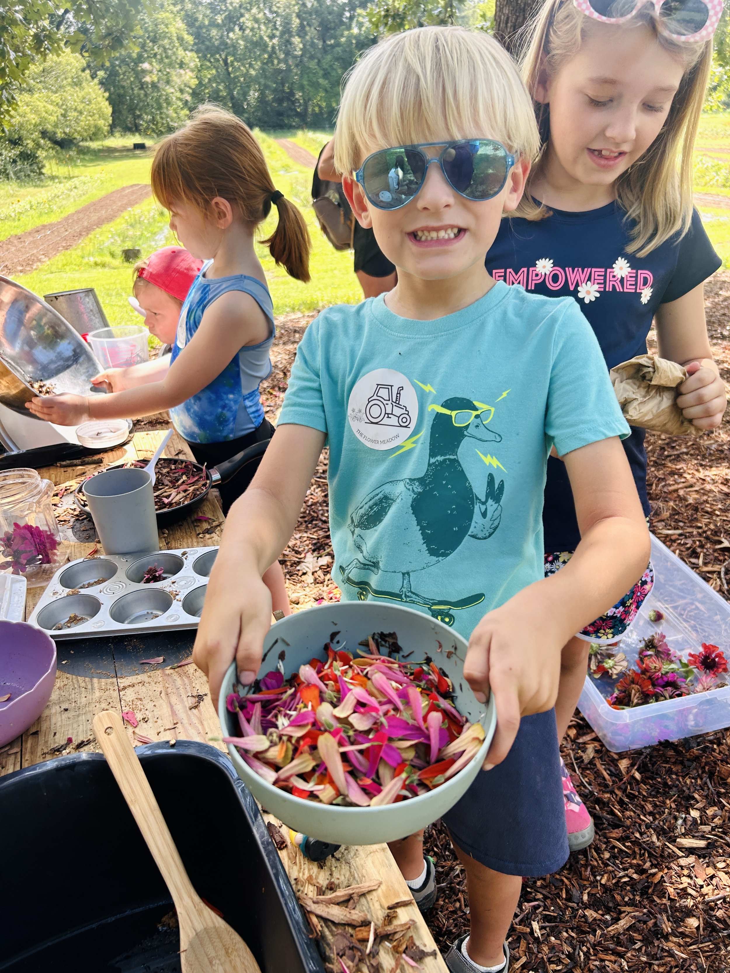 Kids outdoors around a wooden table with flower petals, bowls, and gardening tools, participating in a flower petal craft activity,  at The Flower Meadow Flower Farm in Coweta, Ok.