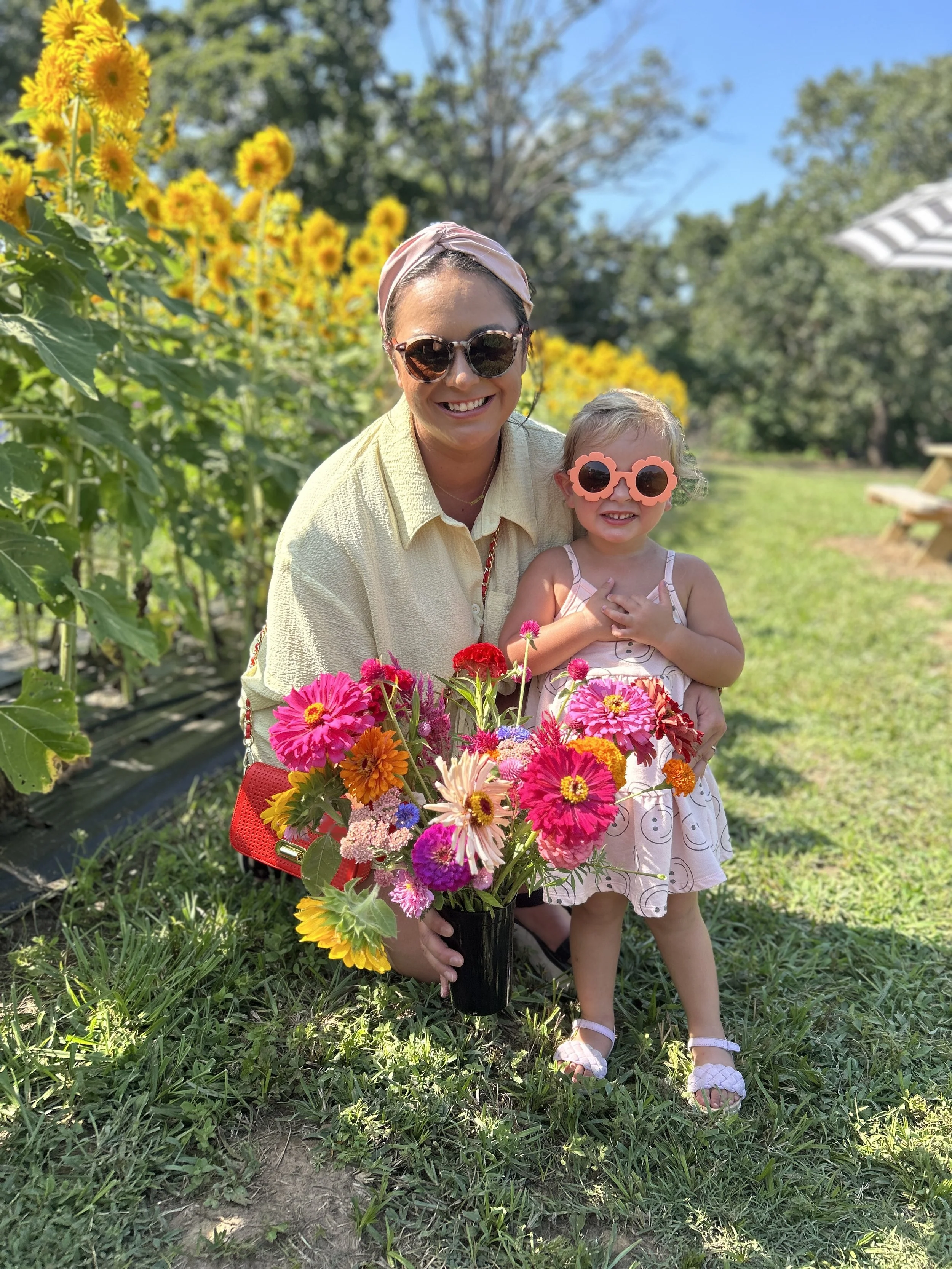 A woman and a young girl smiling outdoors in a garden, holding a colorful bouquet of flowers. They are wearing sunglasses, and the girl is in a light pink dress with a pattern, while the woman is wearing a yellow shirt and a headscarf