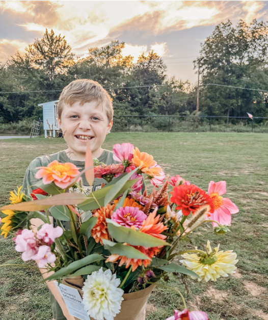 A young boy smiling and holding a large bouquet of colorful flowers outdoors during sunset at The Flower Meadow You Pick Flower Farm and Barnyard Petting in Coweta, Ok