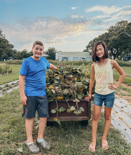 A boy and girl standing next to a garden cart filled with harvested produce, outdoors during late afternoon.