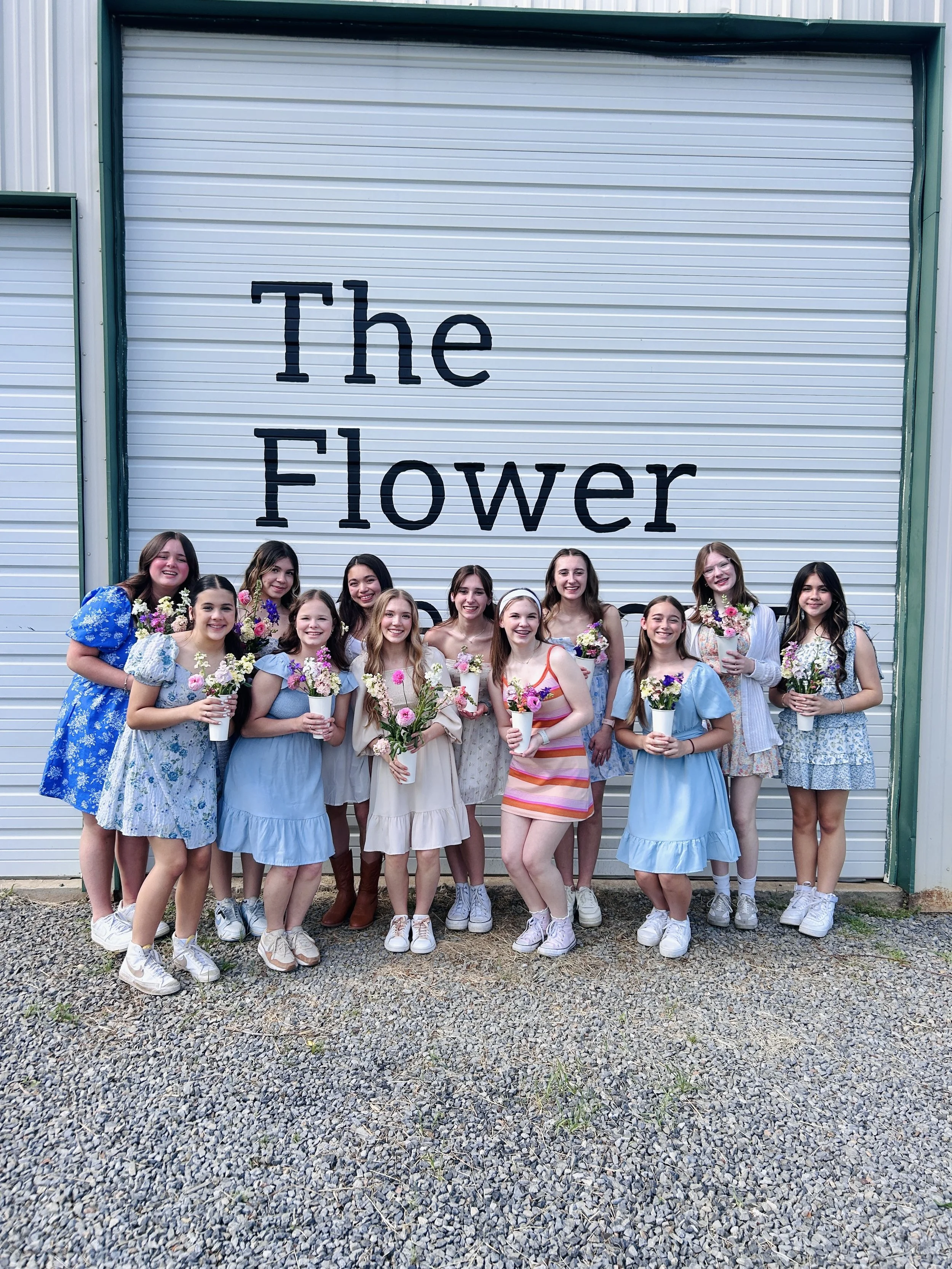 Group of young women holding flowers, standing in front of a sign that reads 'The Flower Meadow. Celebrating a sweet 16.
