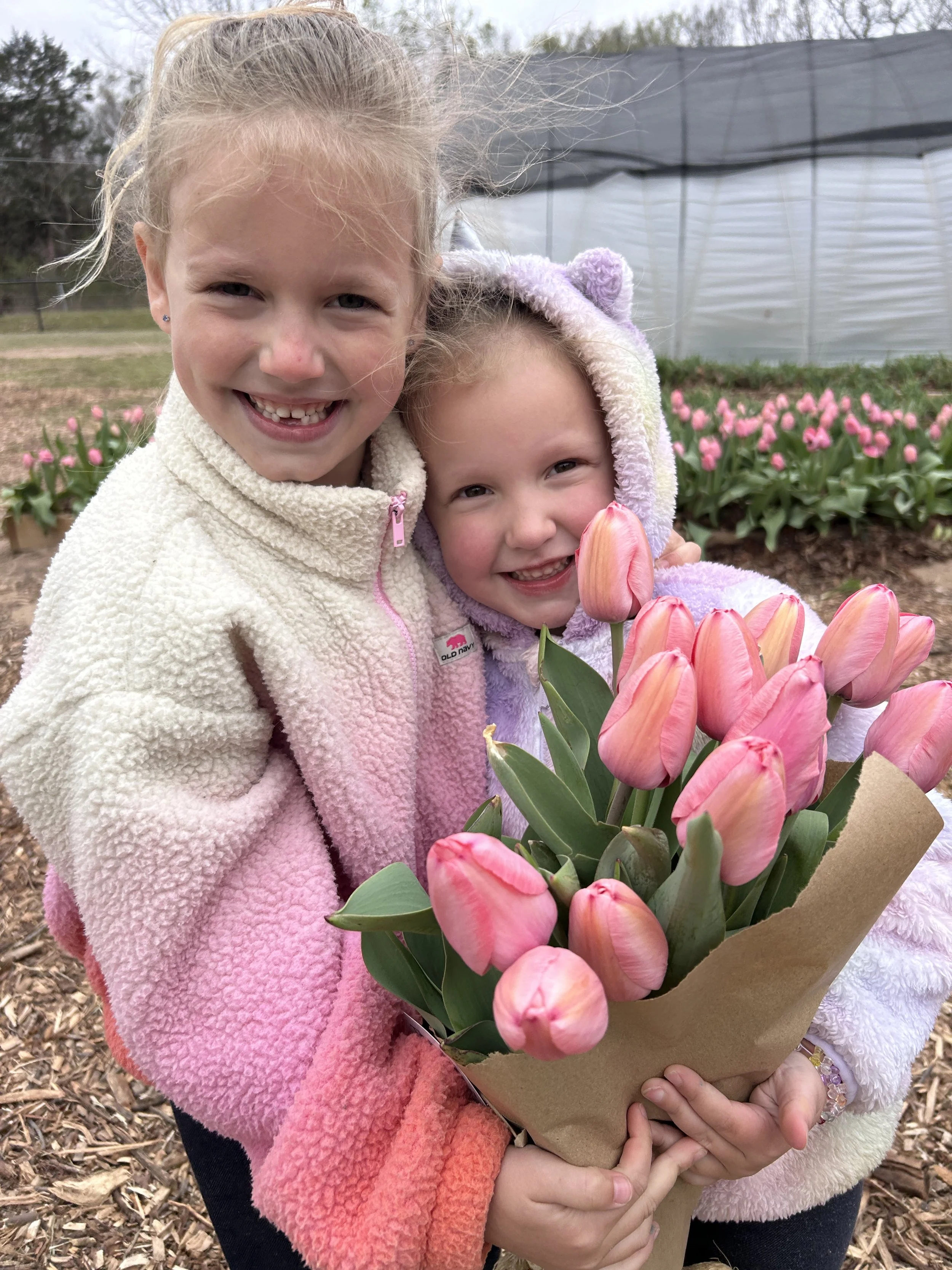 Two young girls holding a bouquet of pink tulips outdoors, with a greenhouse in the background  at The Flower Meadow Flower Farm and Barnyard Petting in Coweta, Ok