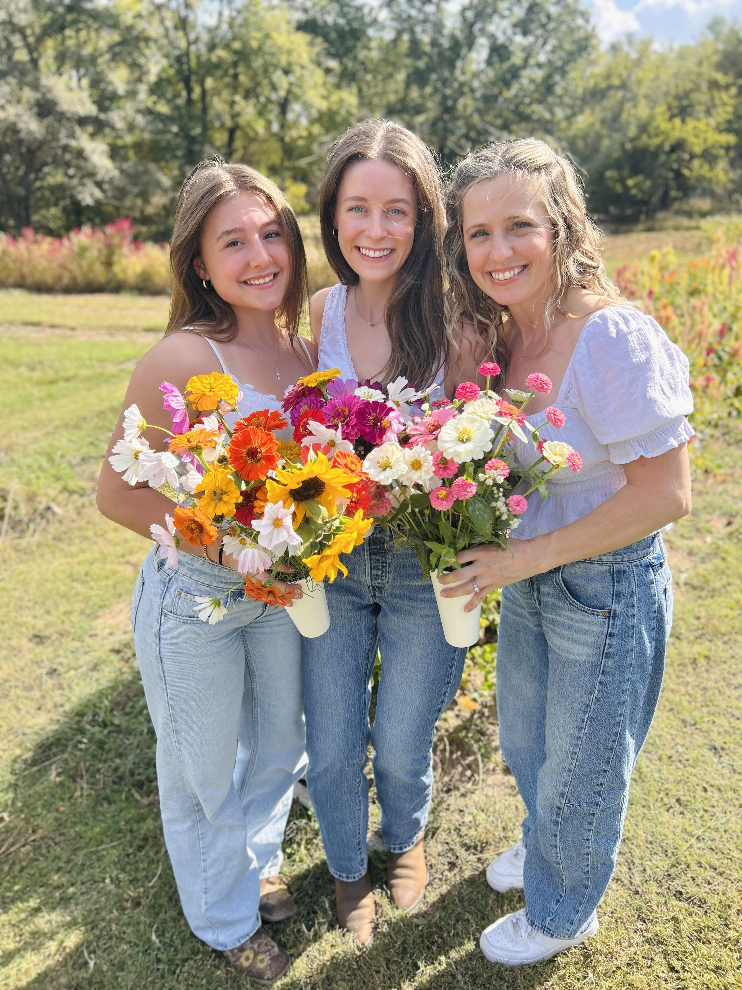 Three women standing outdoors in a park holding colorful flower arrangements in white vases, smiling at the camera with trees and grass in the background on a sunny day. at The Flower Meadow You Pick Flower Farm and Barnyard Petting in Coweta, Ok