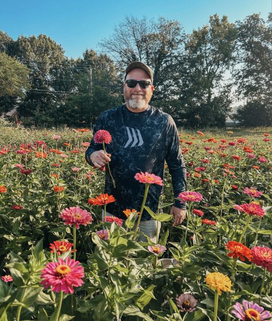 A farmer, wearing sunglasses, a cap, and a dark long-sleeve shirt, standing in a colorful flower field holding a pink flower, with trees and a bright blue sky in the background at The Flower Meadow You Pick Flower Farm and Barnyard Petting in Ok.