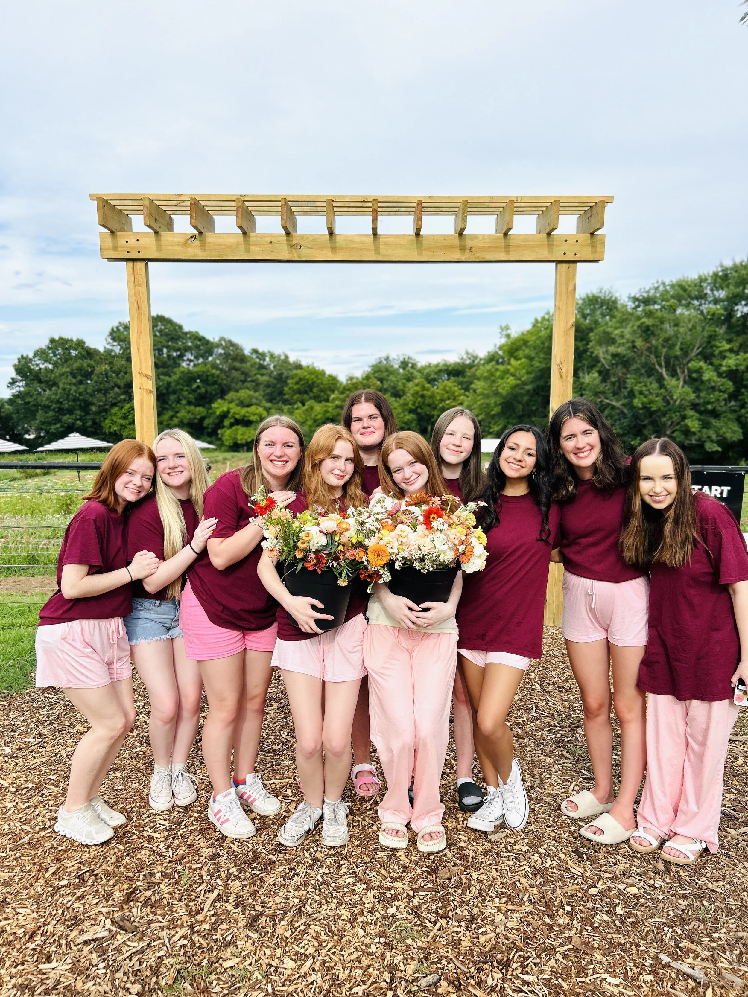 Group of nine young women standing outdoors on a mulched area, smiling, wearing matching maroon shirts and pink pants, a bridal Party picking flowers for the bride's wedding the next day  at The Flower Meadow Flower Farm Coweta, OK