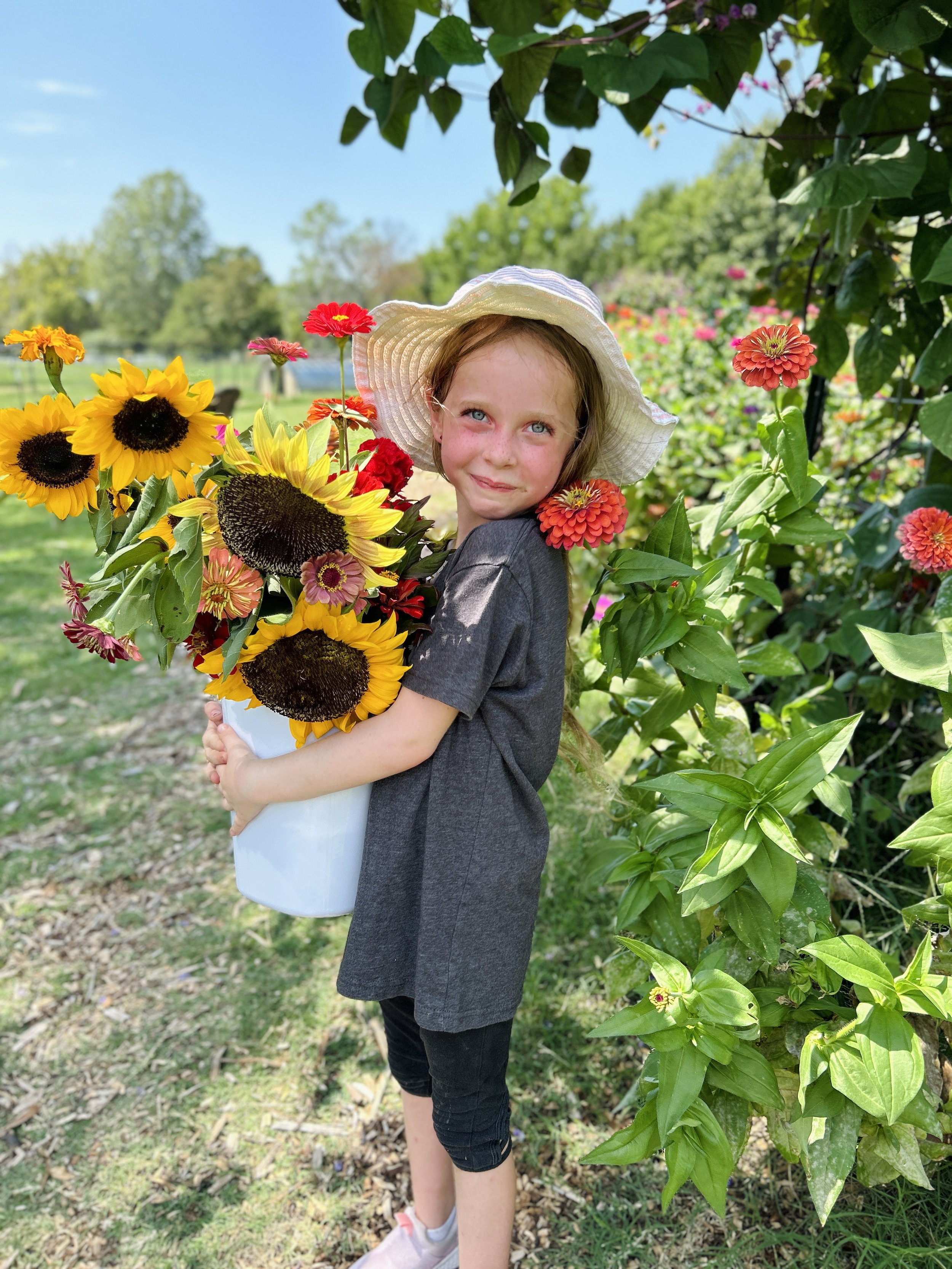 A young girl with light brown hair and blue eyes is holding a large bouquet of colorful flowers, including sunflowers and marigolds, while standing outdoors in a garden with flowering bushes and trees in the background. She is wearing a white wide-brimmed hat, a dark gray shirt, and black shorts