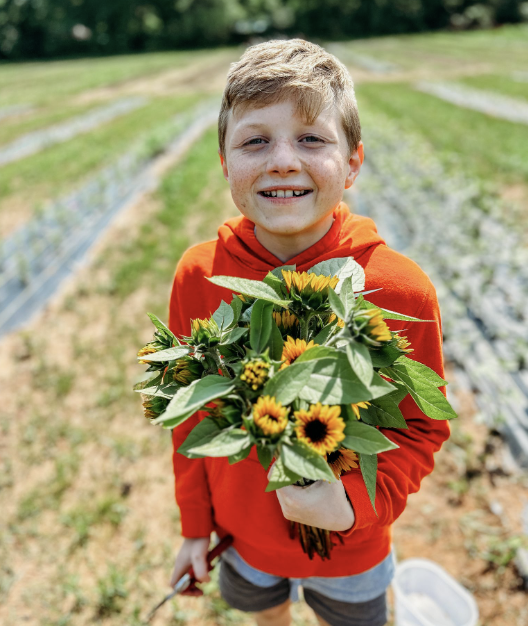 A smiling boy wearing an orange sweatshirt holding a bouquet of sunflowers and greenery in a garden or farm field at The Flower Meadow You Pick Flower Farm and Barnyard Petting in Coweta, Ok