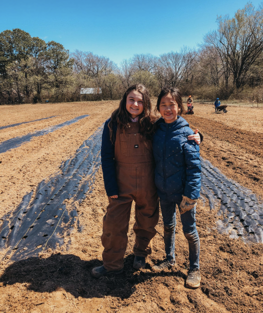 Two young girls smiling, standing close together in a farm field with rows of plastic mulch and trees in the background on a sunny winter day.