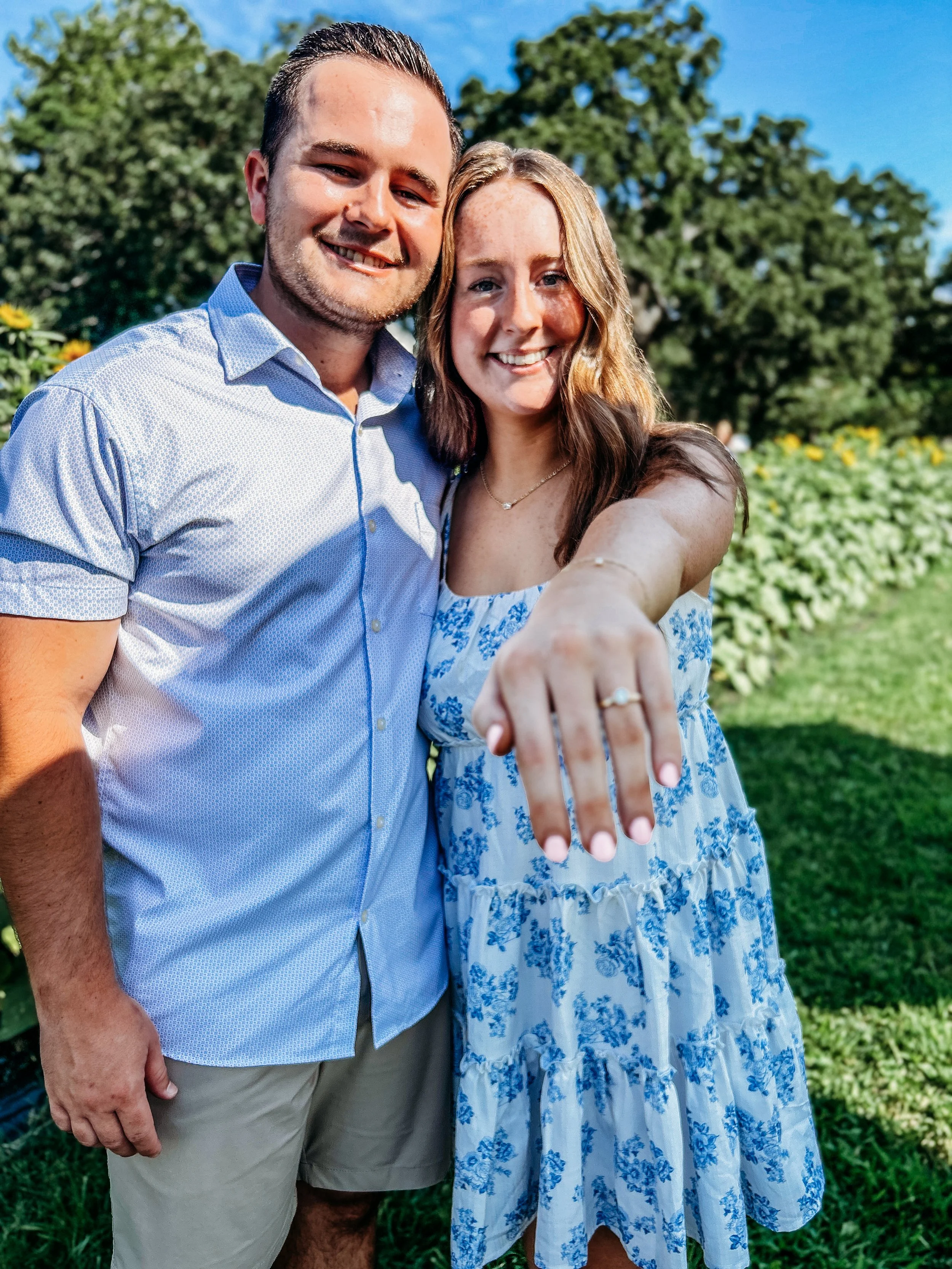 A smiling couple taking a selfie outdoors at The Flower Meadow You Pick Flower Farm and Barnyard Petting in Coweta, Ok celebrating a proposal, the woman showing her wedding ring and hand extended towards the camera.