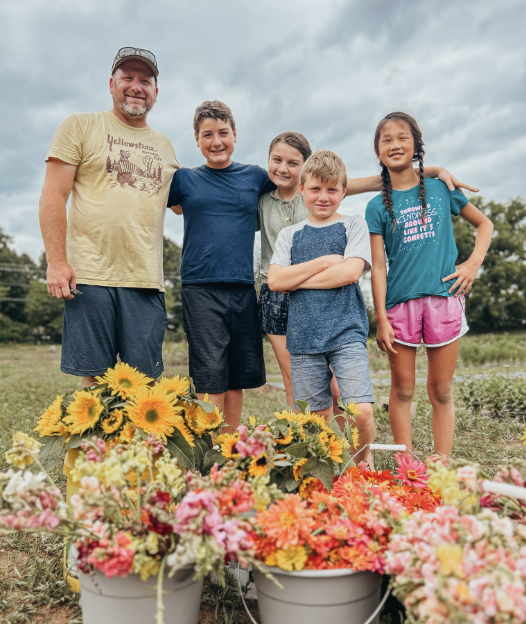 A farm and four children standing outdoors in a row, smiling with arms around each other, in a field with buckets of colorful flowers in the foreground and a cloudy sky overhead.