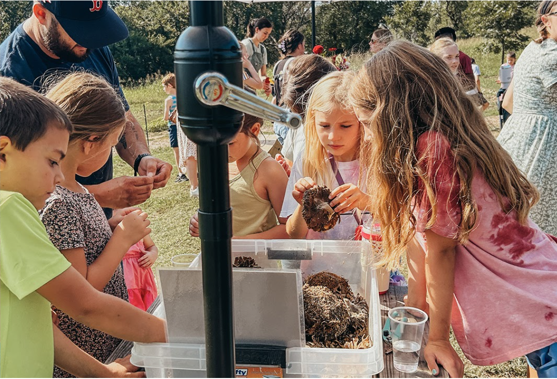 Children gathered around a table outdoors, examining and touching flower seeds and parts as part of a field trip to The Flower Meadow Flower Farm in Coweta, Ok