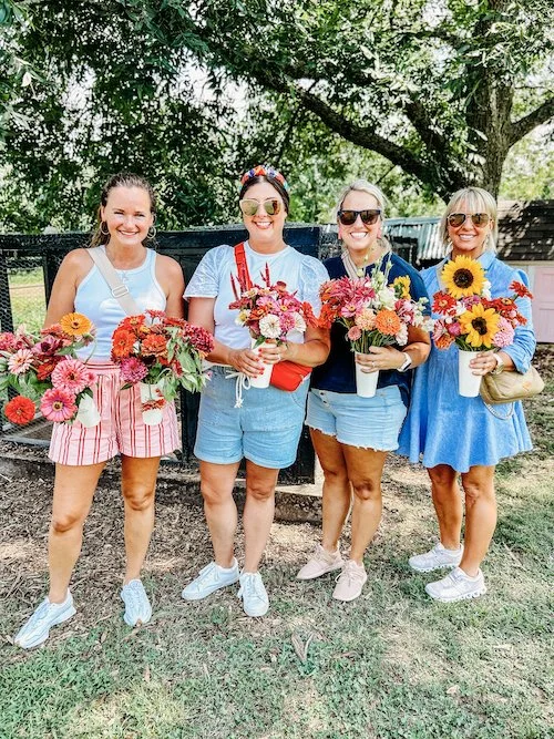 Four women standing outdoors on grass holding vibrant flower bouquets, smiling, with trees in the background at The Flower Meadow You Pick Flower Farm and Barnyard Petting in Coweta, Ok