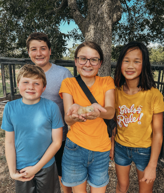 Four children standing outdoors near a tree, smiling. The girl in the center holds a small chick at The Flower Meadow You Pick Flower Farm and Barnyard Petting in Coweta, Ok