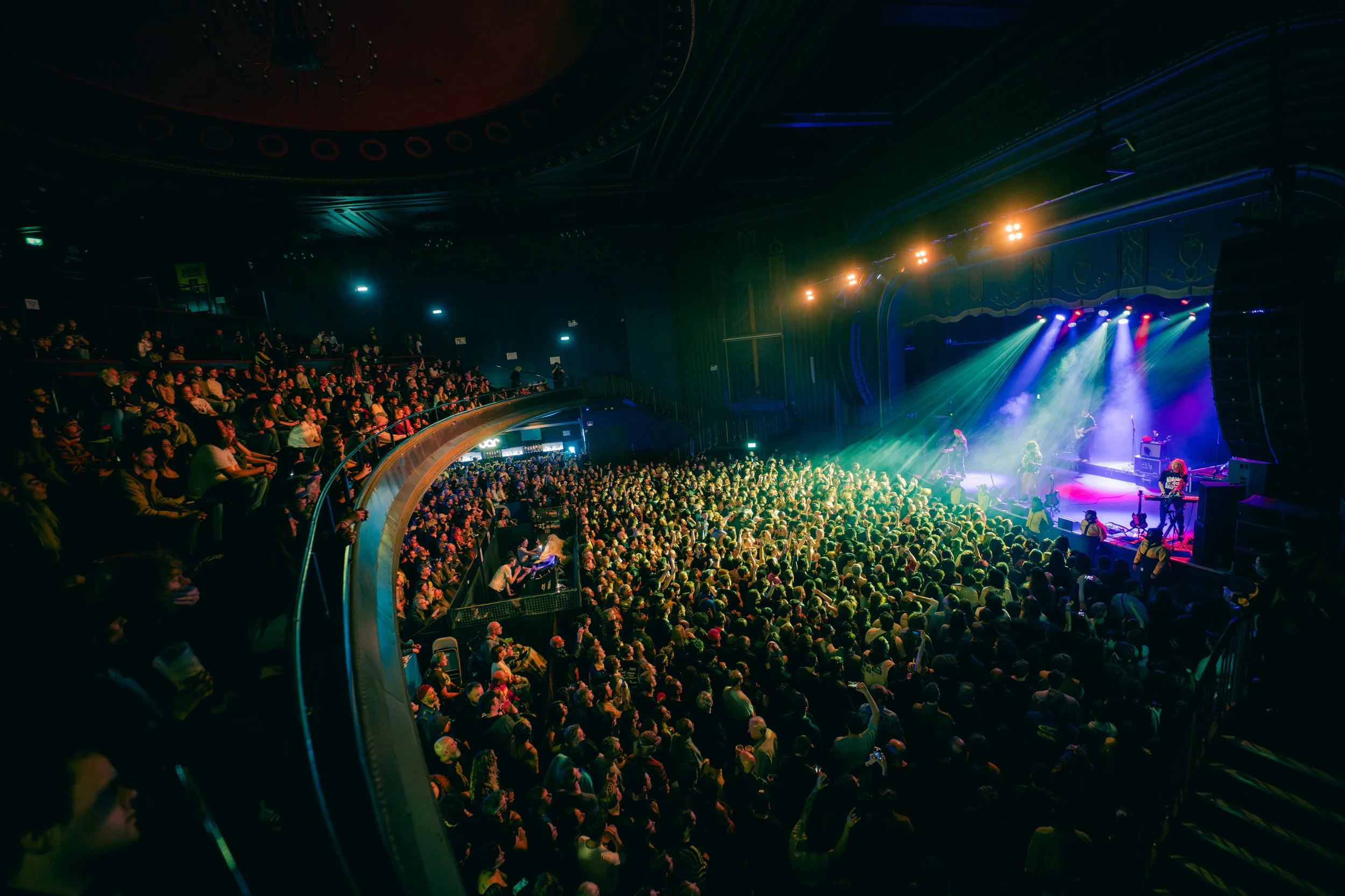 A large crowd watching a live music concert on a stage with colorful lights, inside an indoor venue with multiple seating levels.