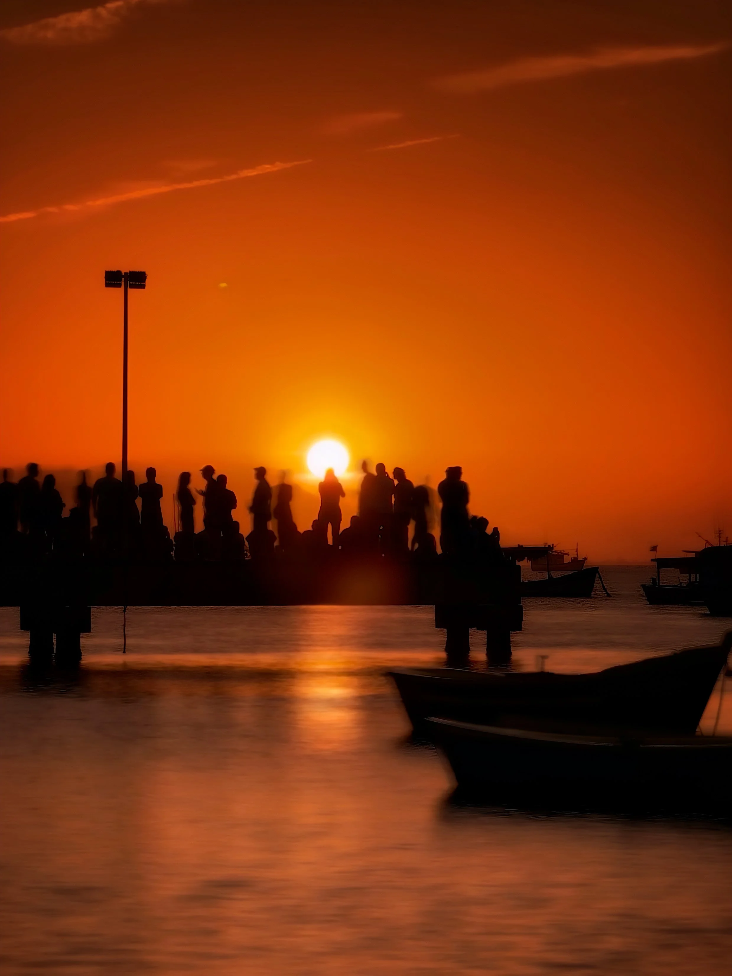 Pessoas silenciosas assistindo o pôr do sol na orla do mar com barcos ao fundo.
