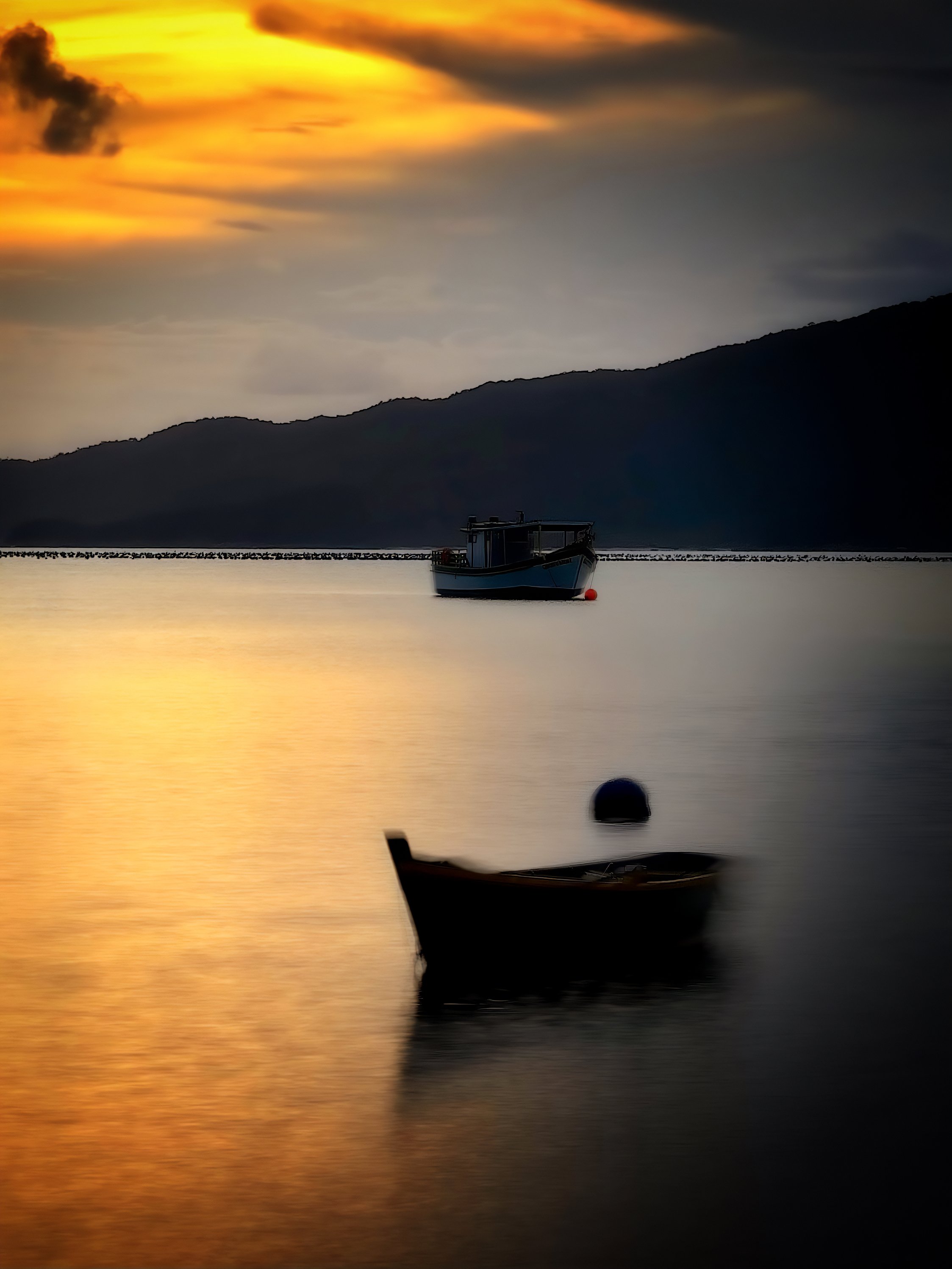 Paisagem de pôr do sol com dois barcos na água tranquila, um em primeiro plano e outro ao fundo, com montanhas ao longe e céu com nuvens douradas.