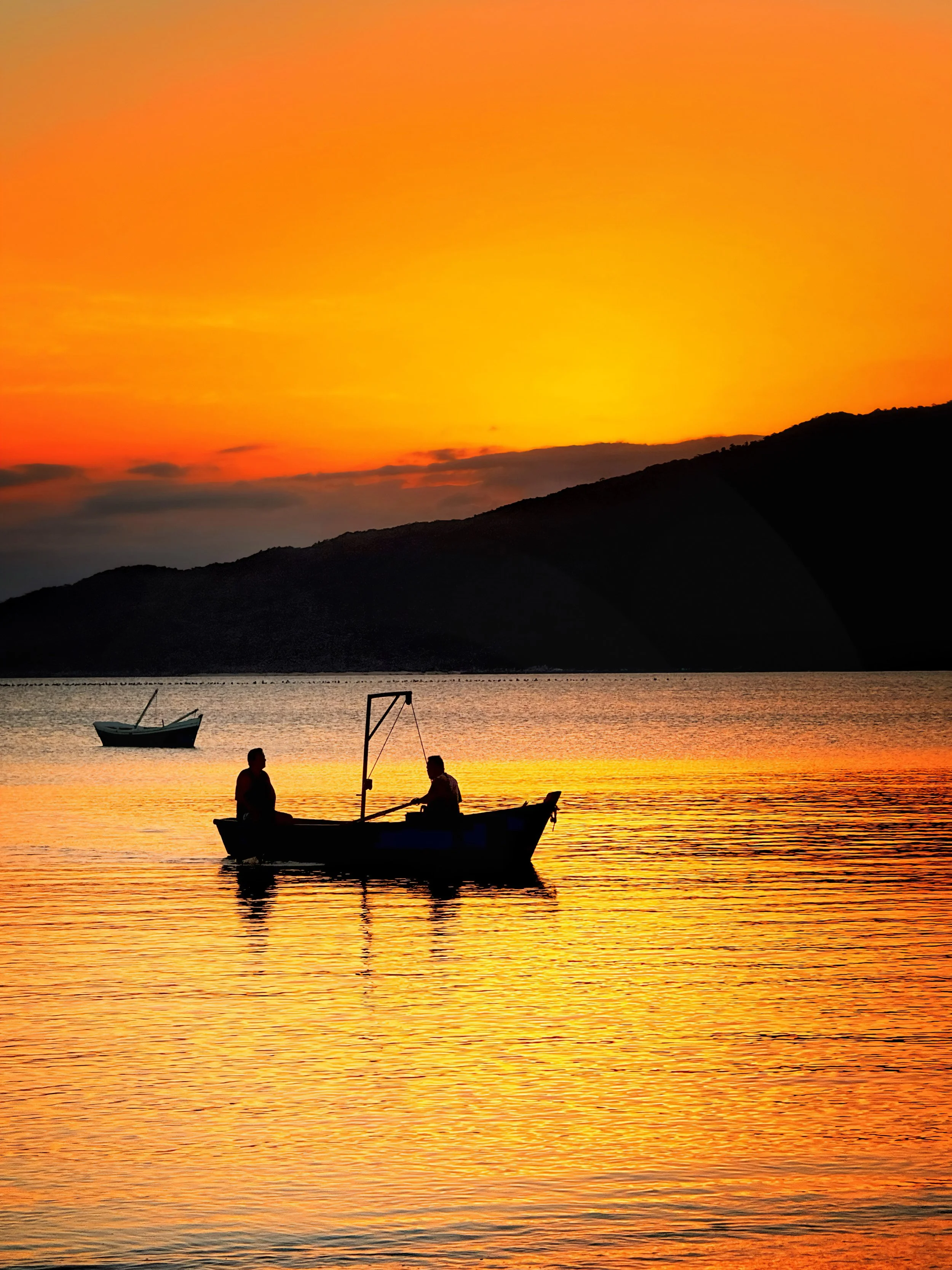 Pôr do sol com céu laranja, silhueta de duas pessoas em um barco na água e uma ilha ao fundo.