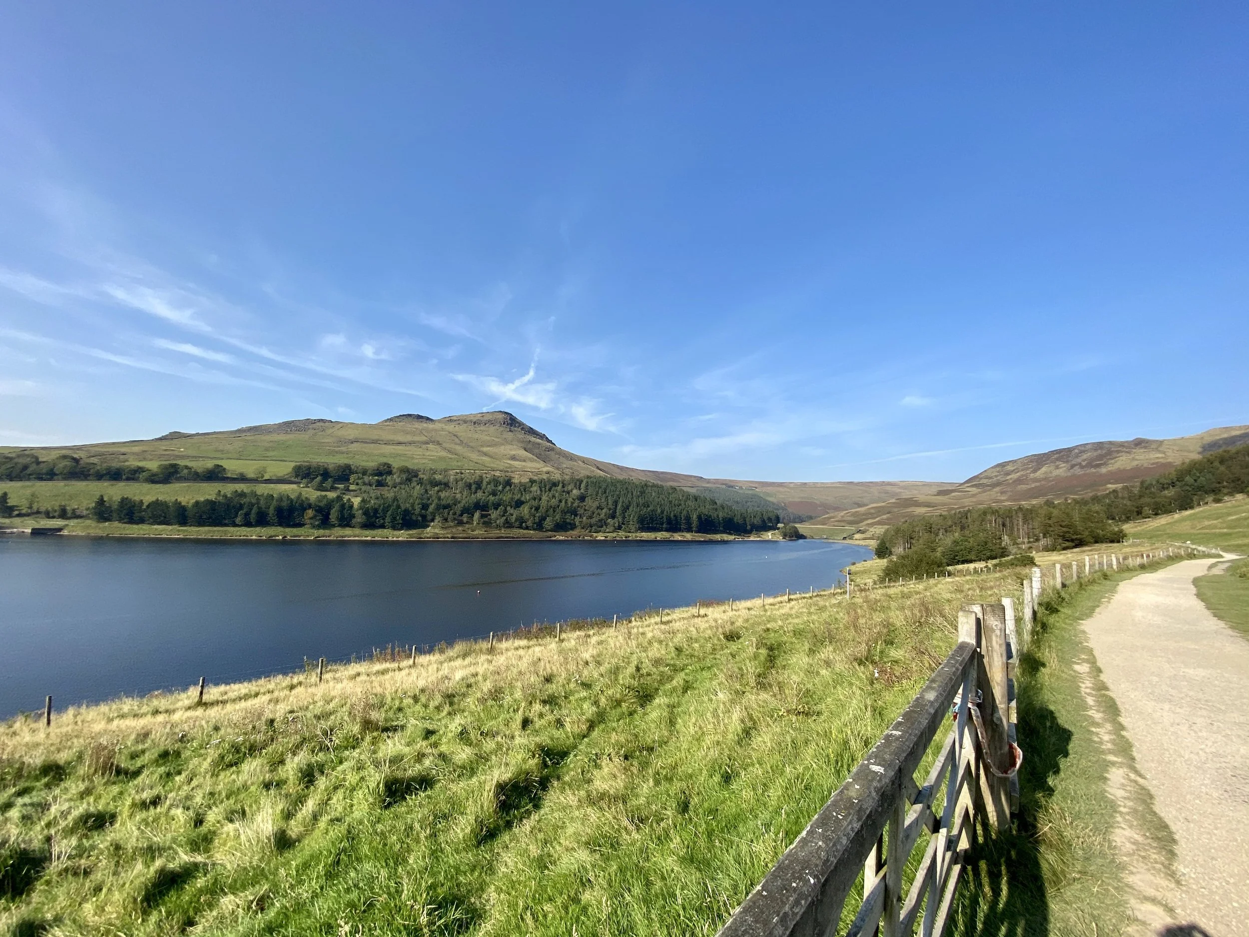 Scenic view of a lake with green hills and mountains in the background under a clear blue sky, with a gravel path and a wooden fence on the right side.