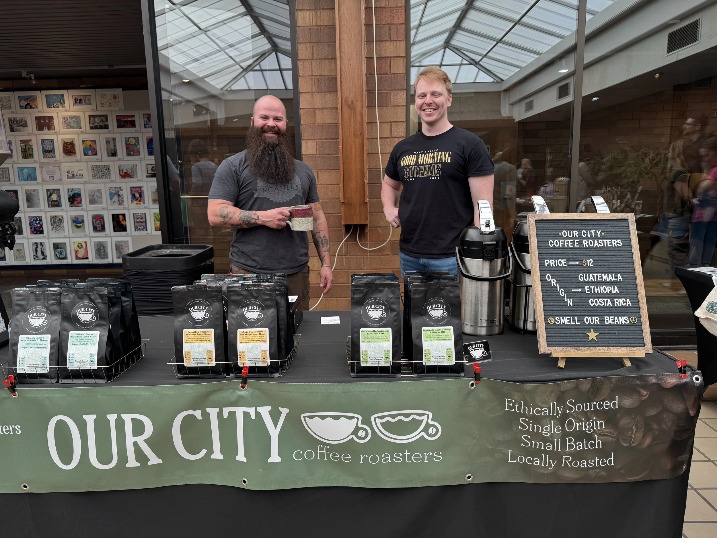 Two men standing behind a table at a coffee booth in a shopping mall. One man has a beard and tattoos, holding a mug; the other is smiling. The table displays bags of coffee and a sign reading "Our City Coffee Roasters." There are people and artwork in the background.