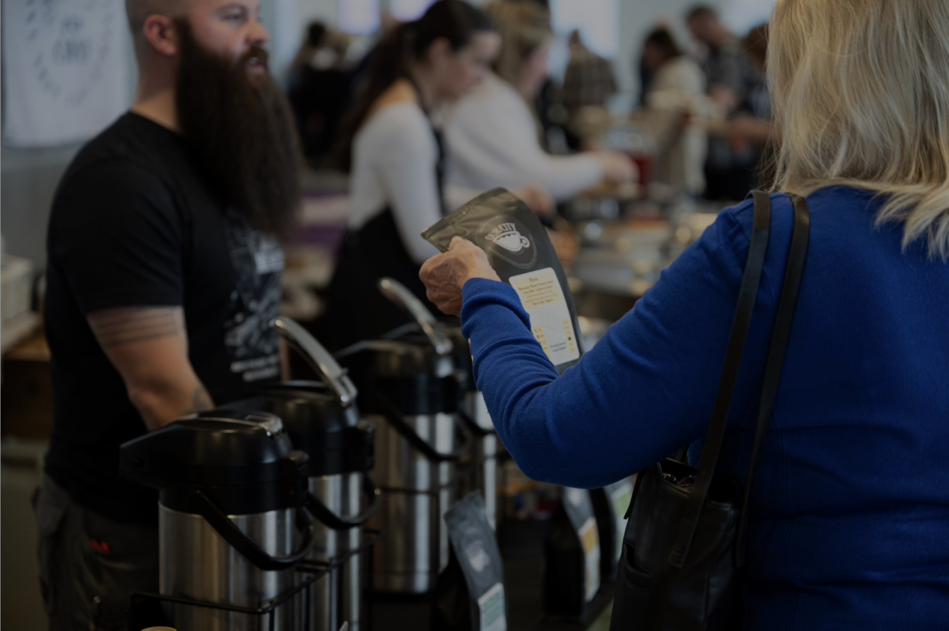 A blonde woman in a blue shirt holding a package, purchasing coffee at a coffee shop counter. A barista, with a beard and a black shirt, stands behind and prepares the order, while other customers wait in the background.