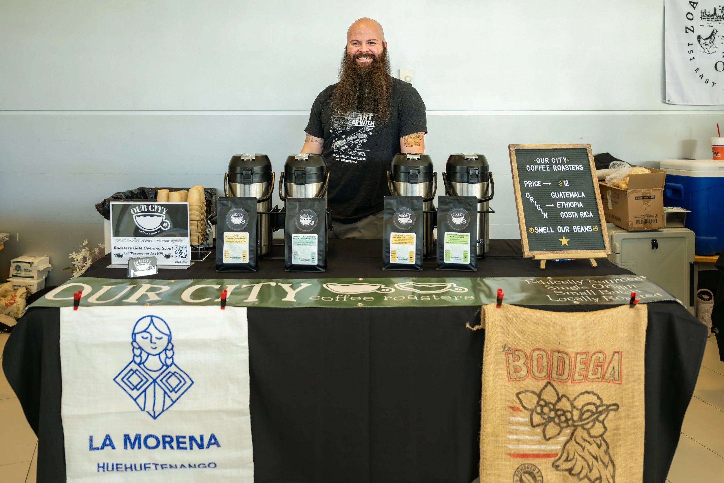 A man with a beard and a black t-shirt standing behind a coffee stand with four coffee dispensers. There are signs indicating the coffee is from Guatemala, Ethiopia, and Costa Rica, and that it costs $12. The background has a white wall with a partial flag or poster on the right.