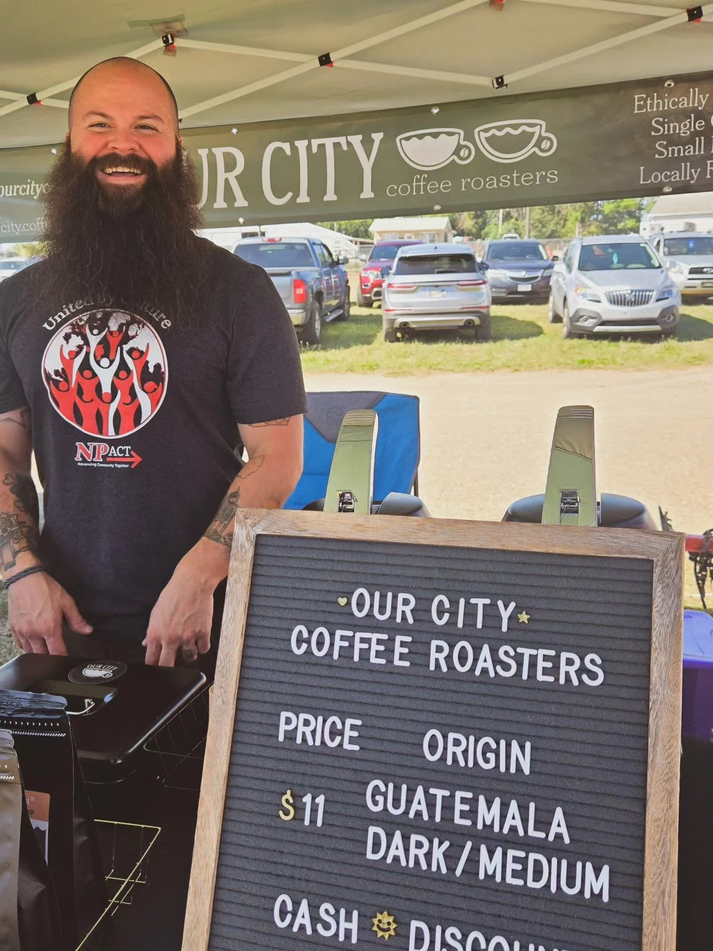 Man with a beard standing behind a signboard at an outdoor coffee stand, with a parking lot in the background.