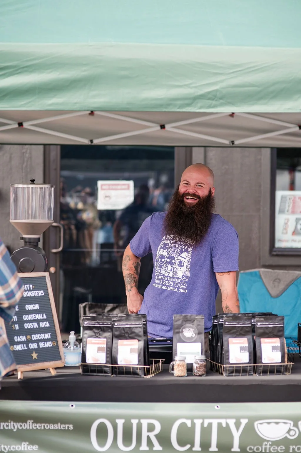 A smiling bearded man at a coffee stand under a green canopy, with coffee bags and jars on the table, at an outdoor market.