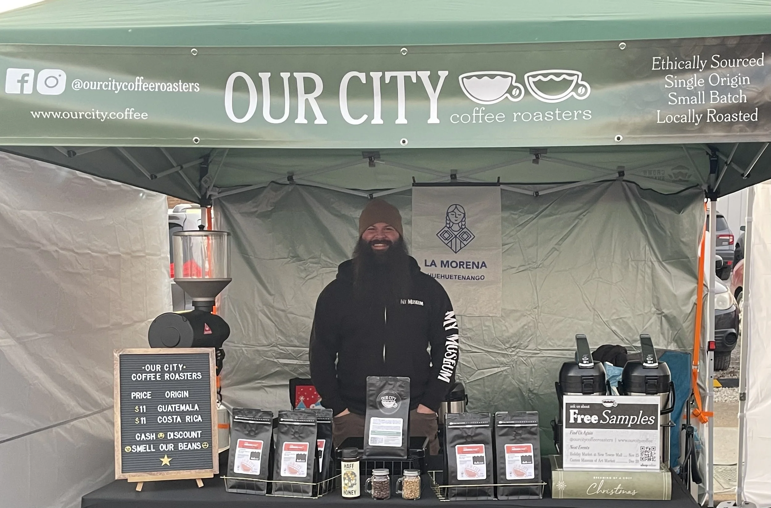A man with a beard and beanie standing behind a coffee stand with a green canopy that reads "OUR CITY coffee roasters." The stand has various coffee bags, a sign with pricing from Guatemala, Costa Rica, and Belize, and a small sign advertising free samples. The background shows parked cars and part of the street.