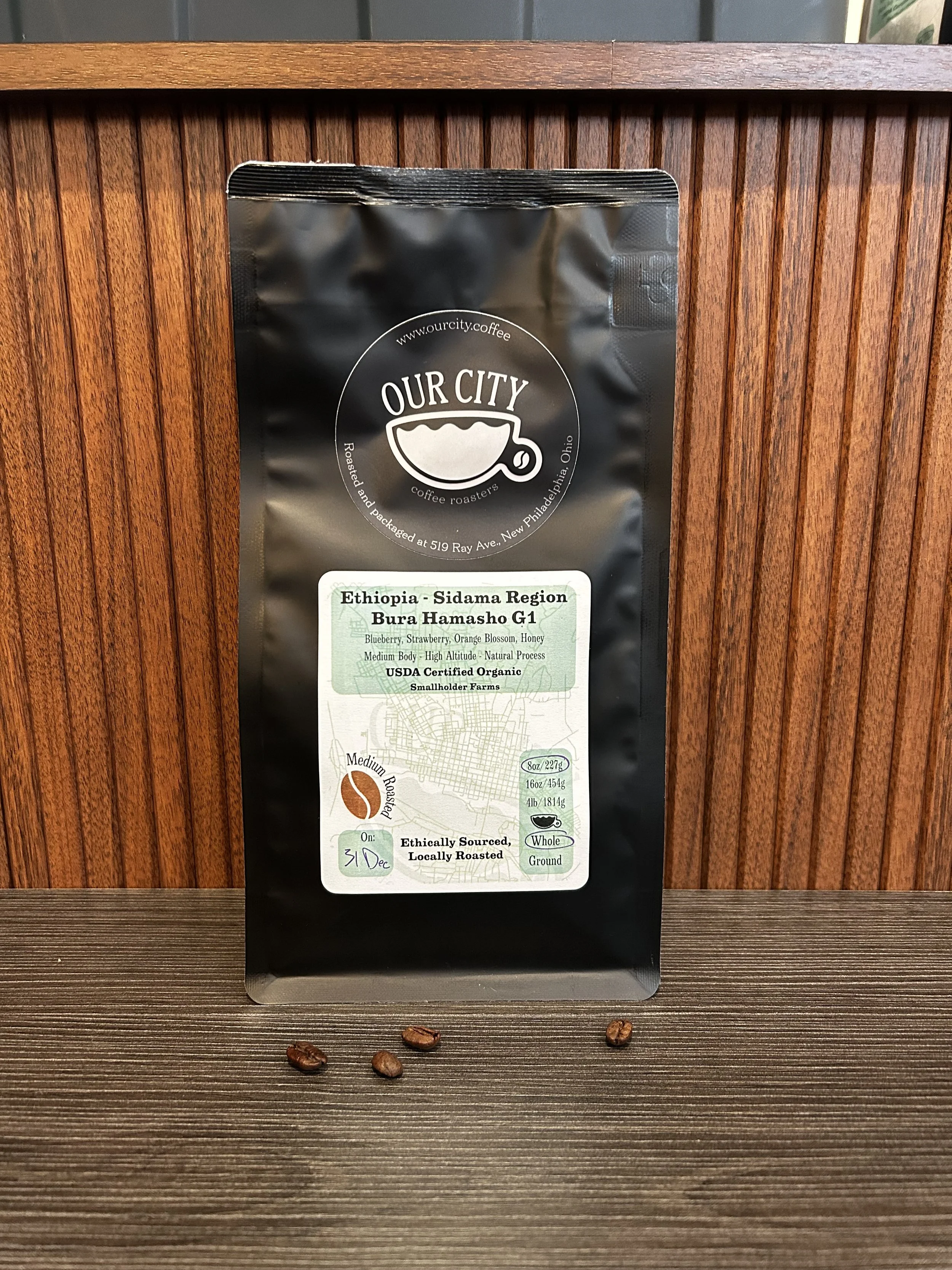 A black coffee bag from Our City Coffee, placed on a wooden surface with some roasted coffee beans underneath. The bag indicates the coffee's origin from Ethiopia, Sidama Region, Bura Hamasho G1, and details about flavor notes and organic certification.
