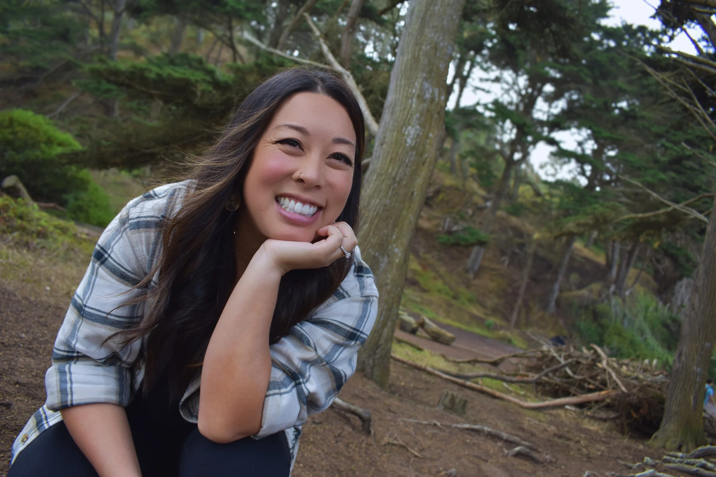 Smiling woman with long dark hair, wearing a plaid shirt, posing outdoors in a wooded area.