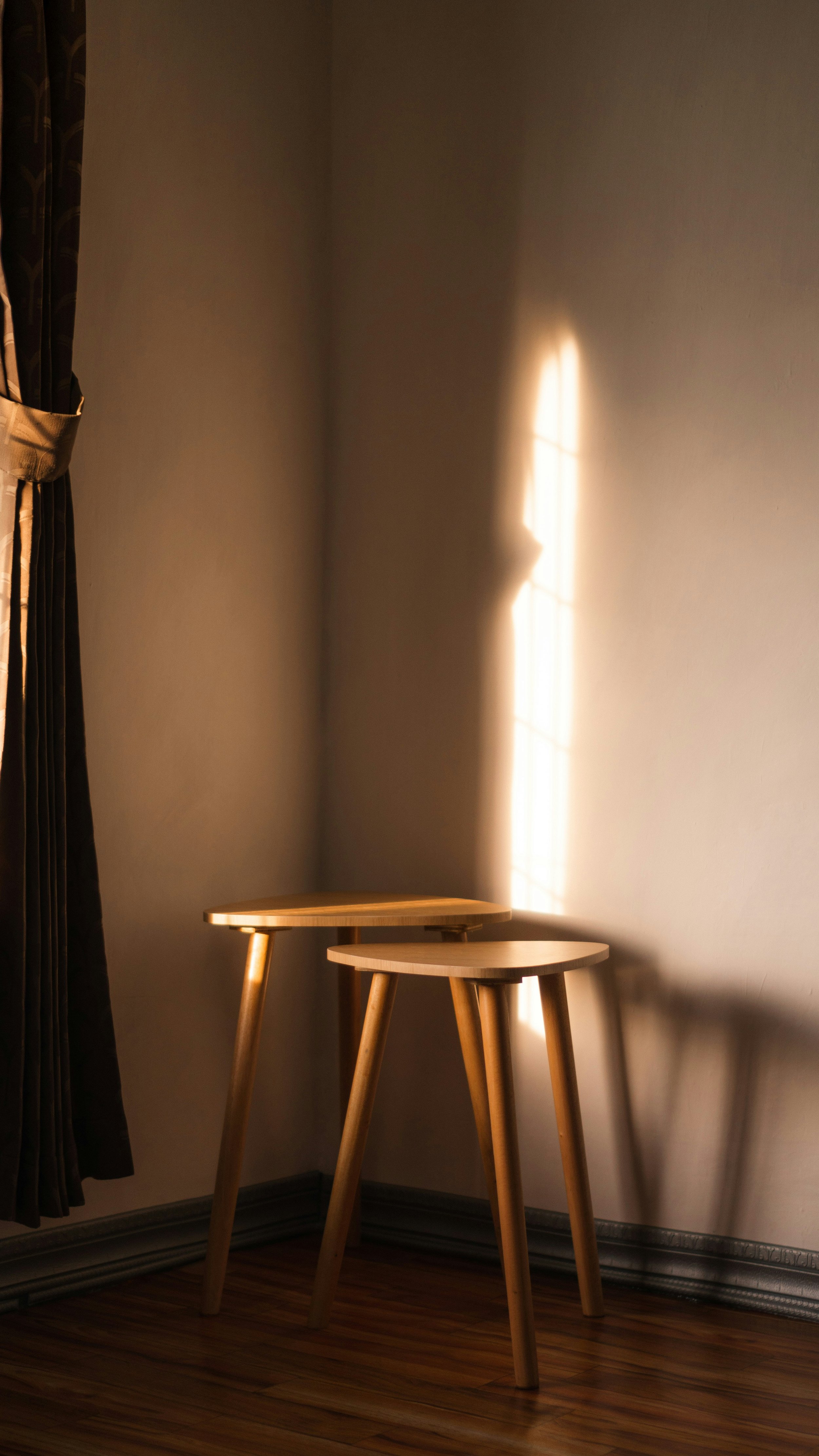 Deux tabourets en bois clair dans un coin d'une pièce, avec un mur beige clair et une fenêtre laissant entrer la lumière.