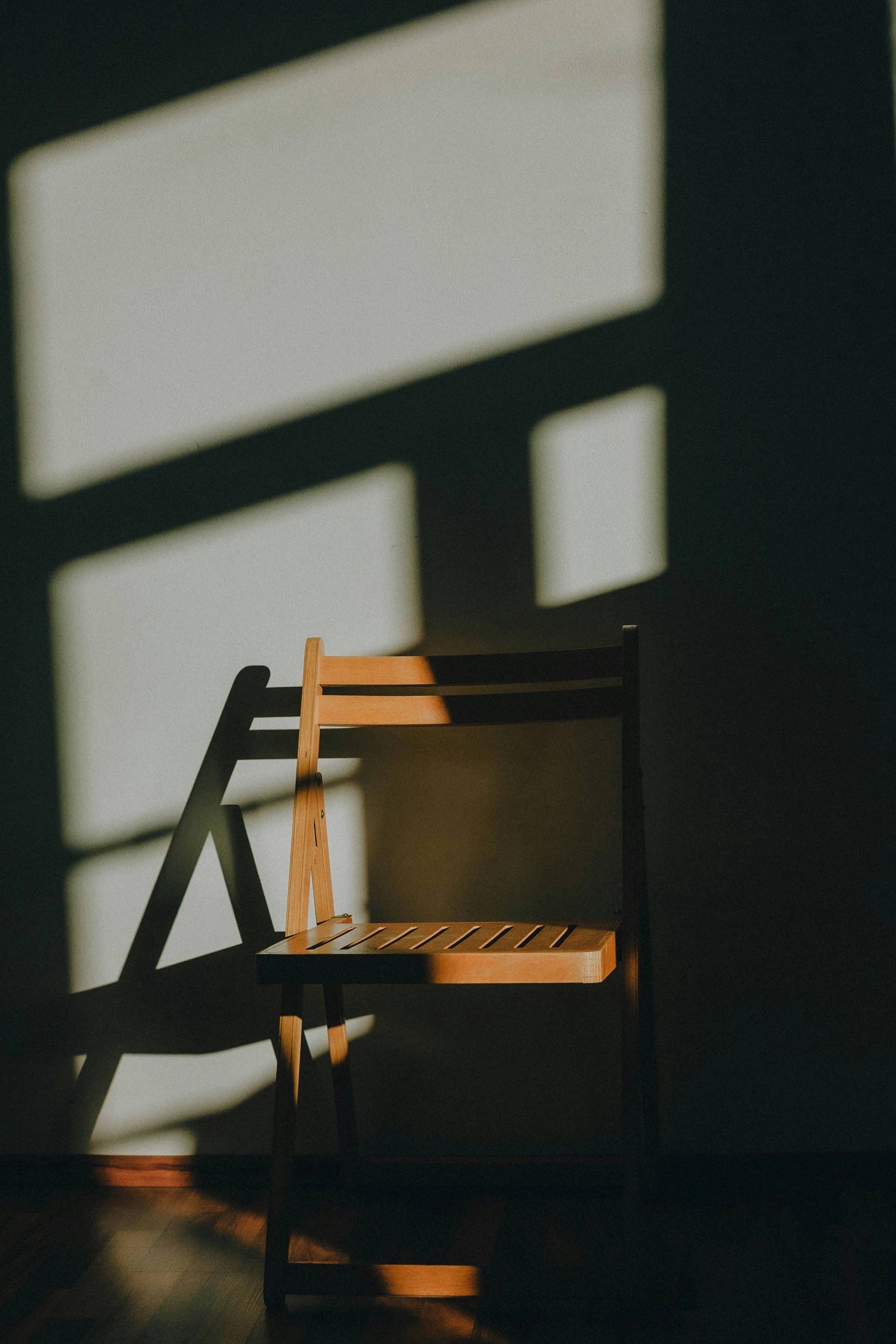 Une chaise en bois dans une pièce sombre, éclairée par la lumière du soleil qui projette des ombres rectangulaires sur le mur.
