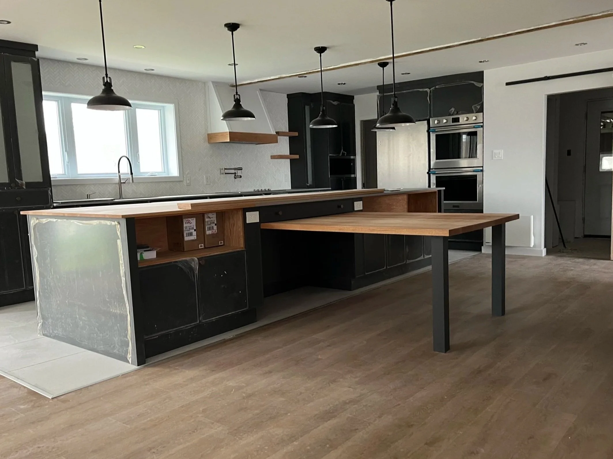 Cuisine moderne avec un plan de travail en bois, des lampes suspendues noires et des armoires noires, fenêtre avec vue extérieure.