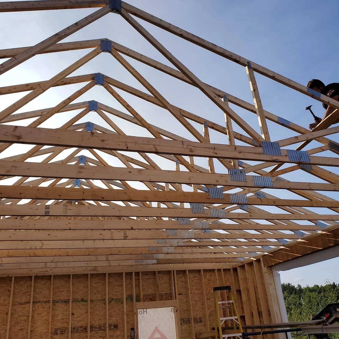 Structure en bois en cours de construction avec un toit en charpente triangulaire, un travailleur utilisant un marteau sur la plateforme supérieure, et un escabeau à l'intérieur du bâtiment.