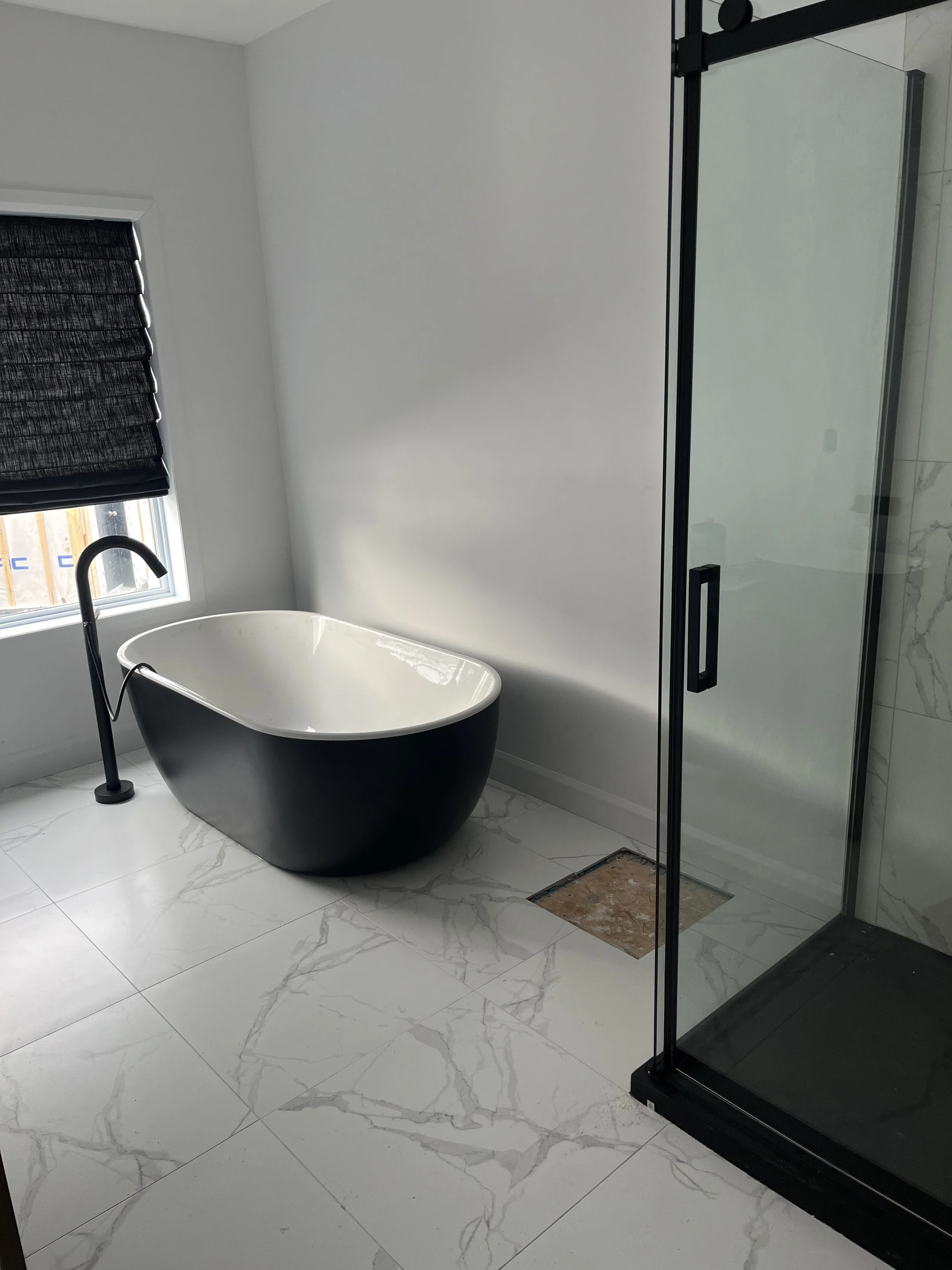 Salle de bain moderne avec une baignoire noire et blanche, une cabine de douche en verre, une fenêtre avec un volet noir, et un sol en carrelage blanc avec des veines grises.