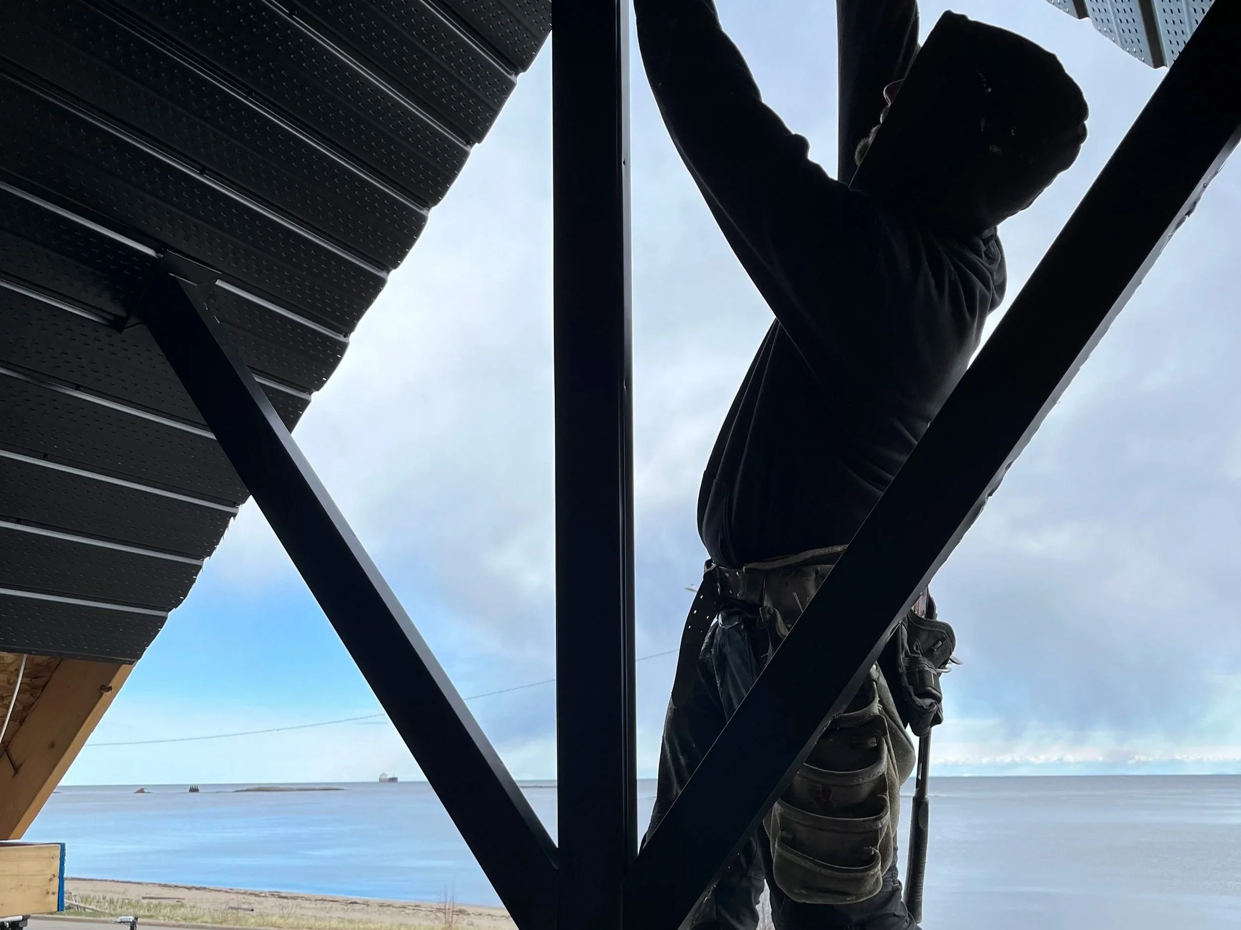 Un homme en noir, portant un masque, grimpe une structure en métal, avec vue sur la mer et le ciel nuageux en arrière-plan.