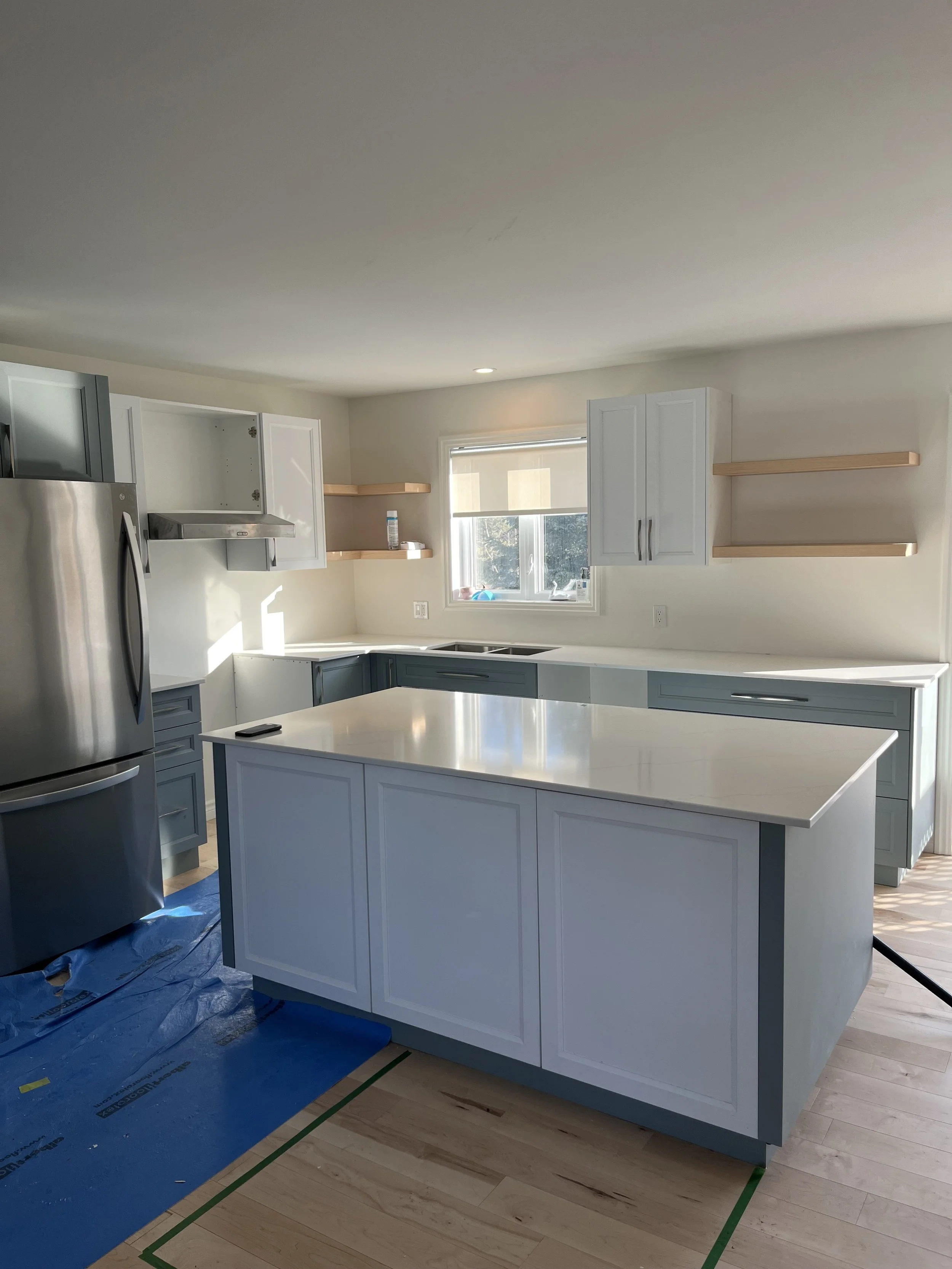 Cuisine moderne avec îlot central, meubles gris, plan de travail blanc, armoires blanches, étagères en bois, fenêtres lumineuses, électroménagers en acier inoxydable.