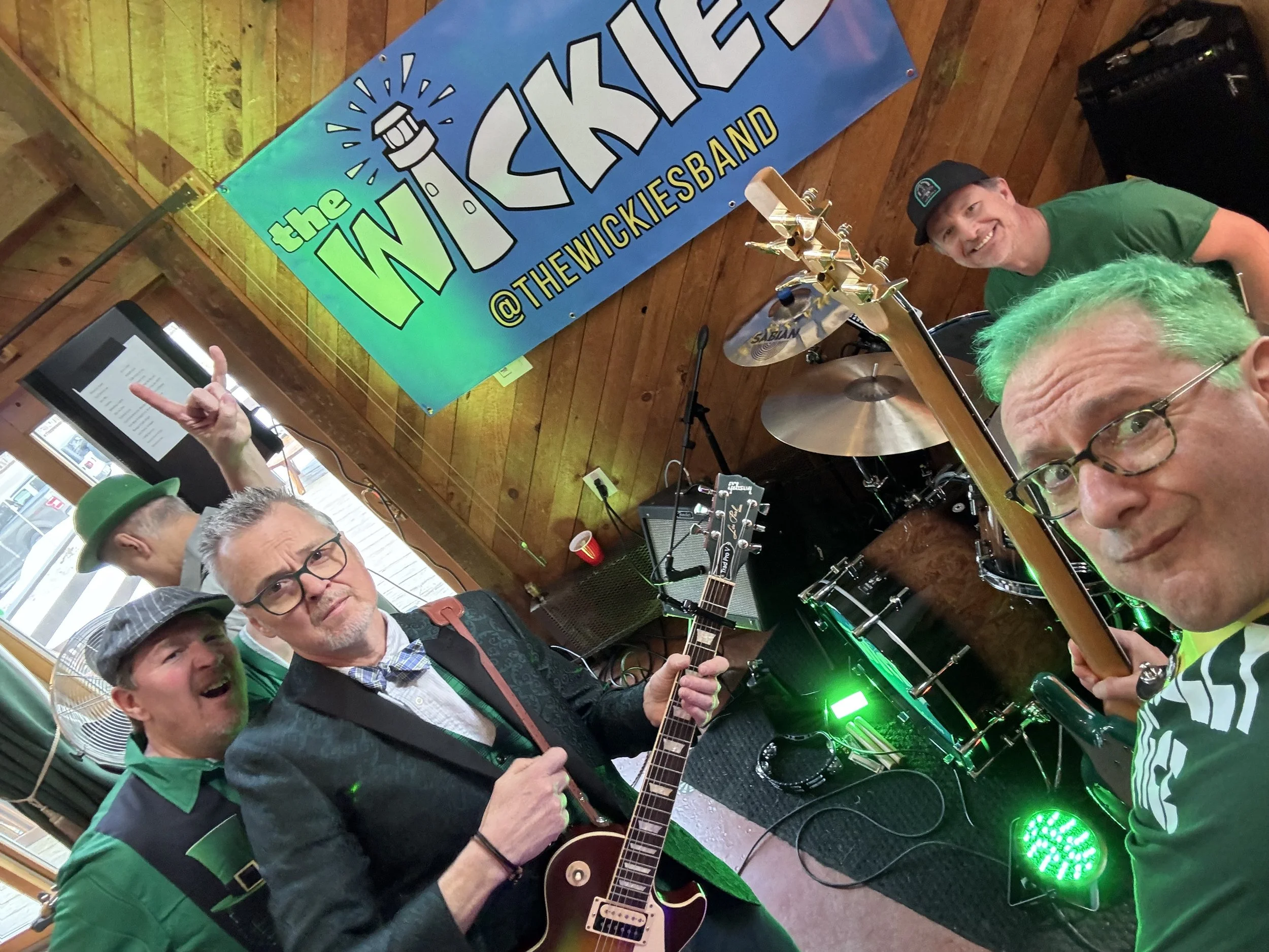 Group of five musicians posing with instruments at a live music event, with a colorful sign in the background that reads 'the WICKIES' at THEWICKIESBAND.