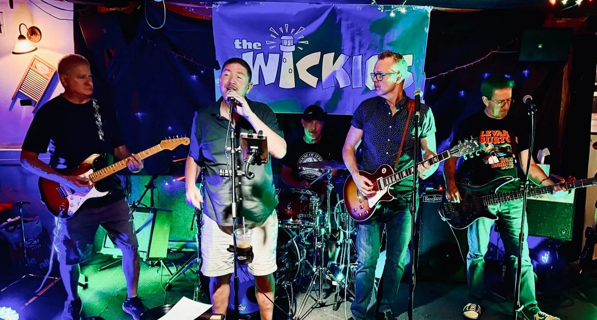A band performing live on stage at a venue with a sign that says 'The WICKIES' in the background. The band has five members playing guitar, drums, and singing, with colorful stage lights and a dark backdrop.