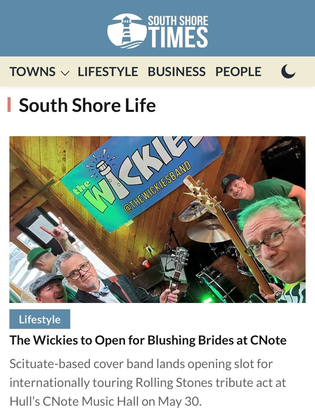 A group of men, one with bright green hair and glasses and holding a guitar, posing with others in front of a colorful banner saying "The Wickies @THEWICKIESBAND" inside a wooden venue. The scene is lively with musical instruments and equipment.