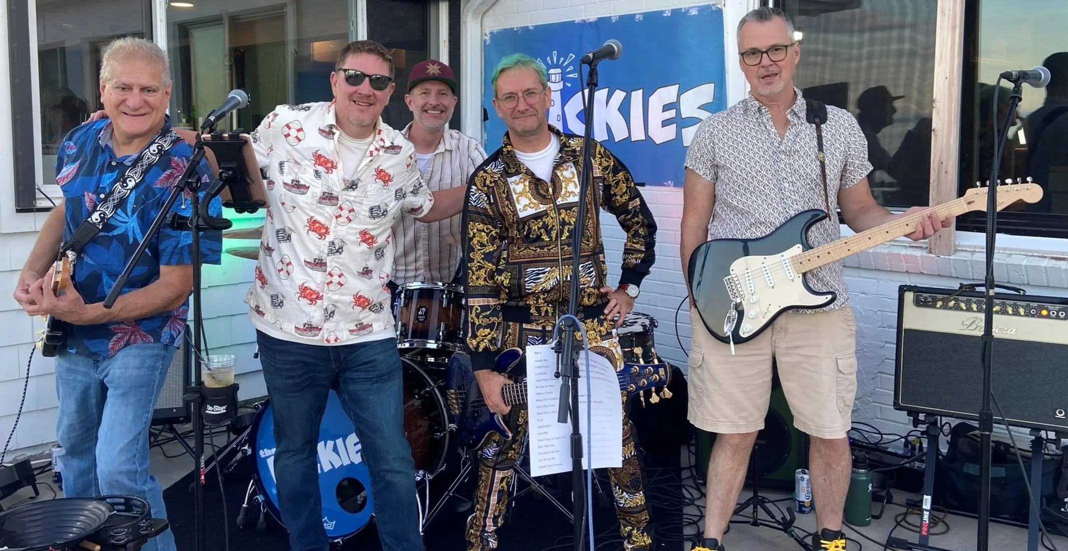 Six men in casual attire with musical instruments, posing outdoors in front of a band performance setup, with a blue banner reading 'the Zookies' behind them.