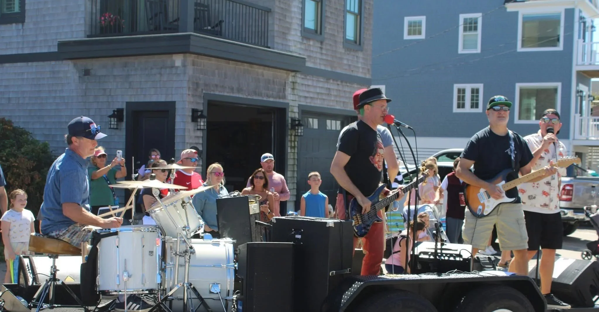A band performing live on a mobile stage outdoors with houses in the background. The band includes a drummer, a vocalist with a guitar, and a guitarist, with an audience watching.